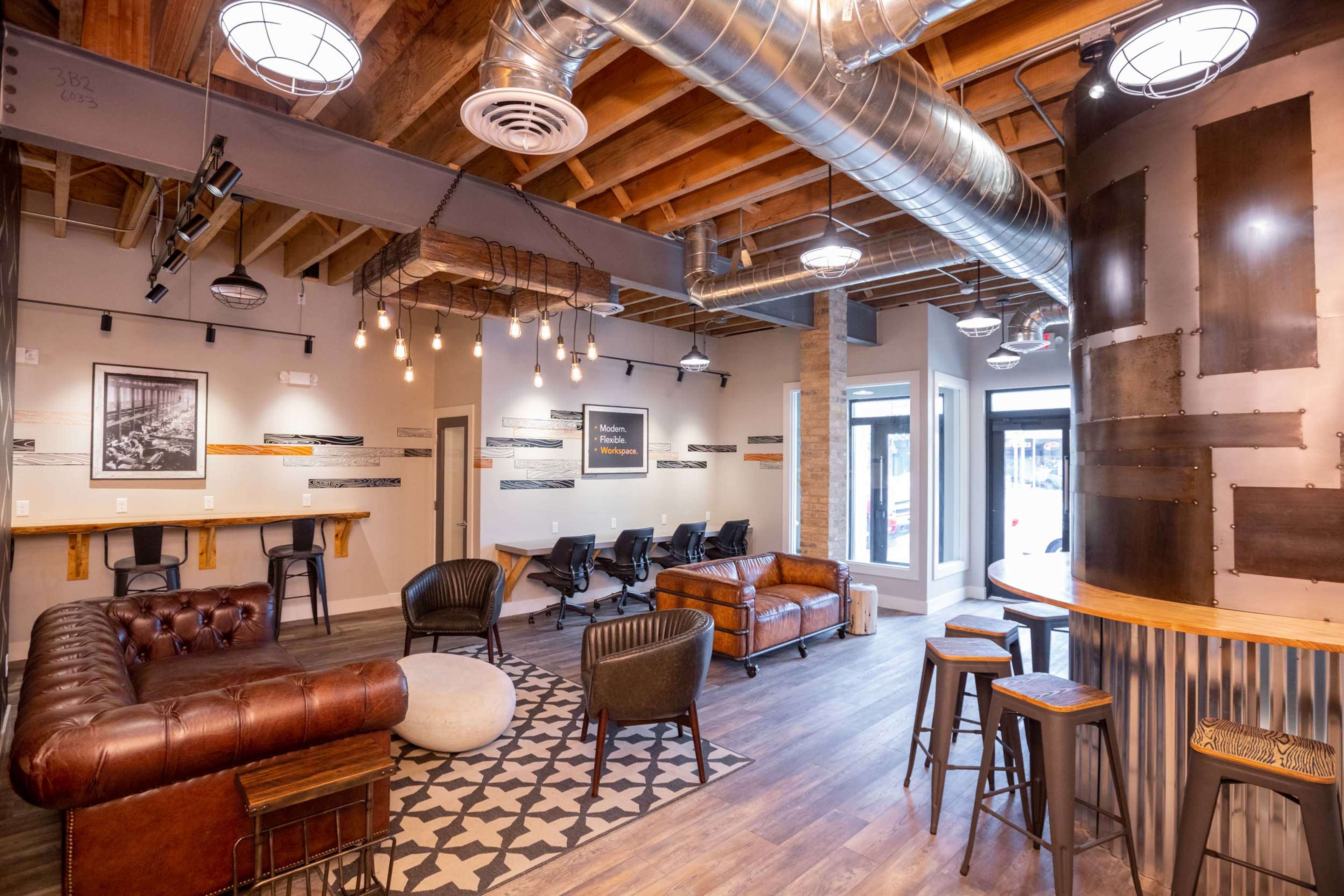 The image shows a modern co-working space featuring a mix of seating arrangements, including a leather sofa, chairs, and bar stools, with large windows and industrial-style lighting.