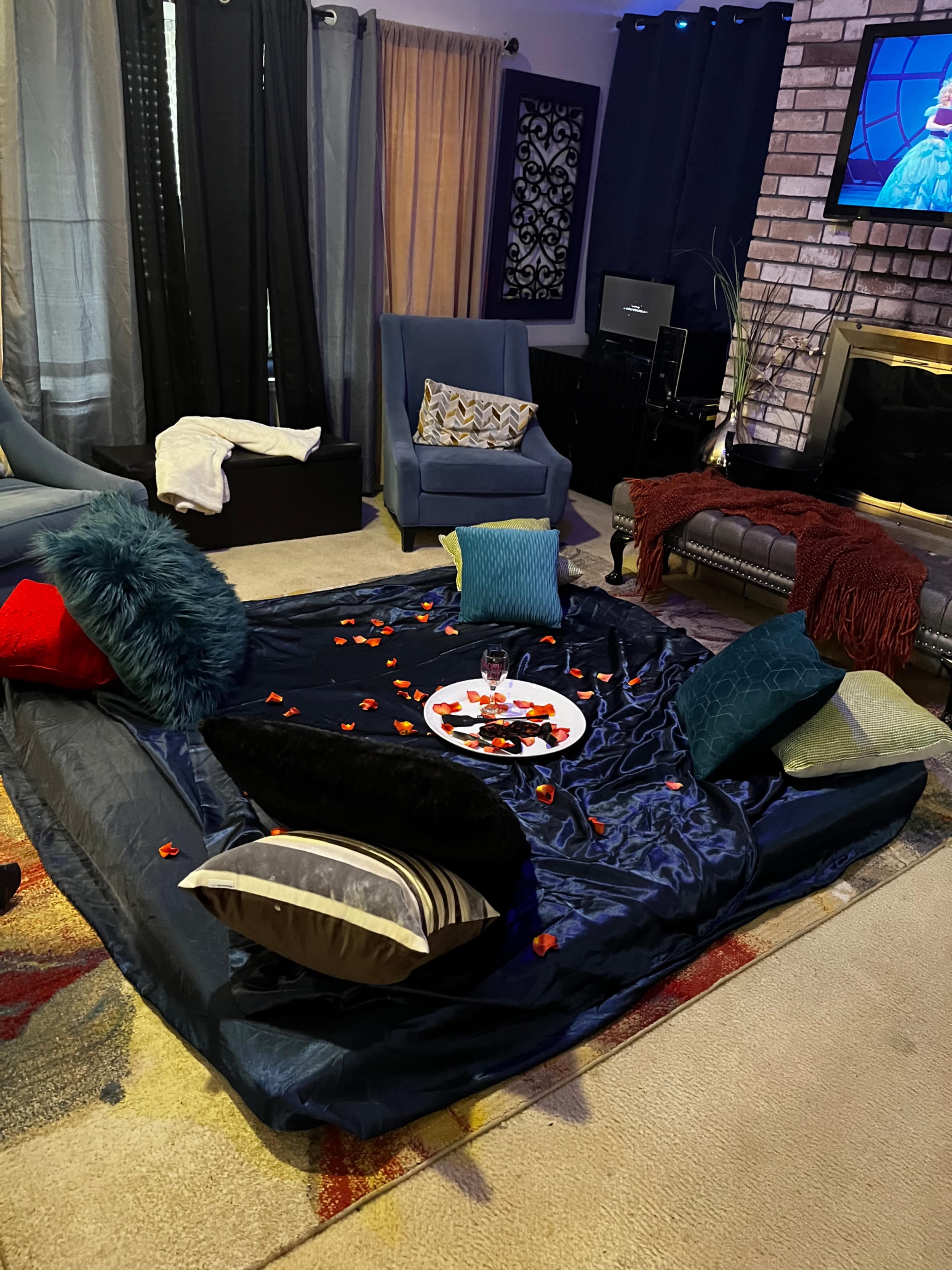 A living room setup with a large dark blanket on the floor surrounded by several cushions and a circular plate in the center, scattered with rose petals.