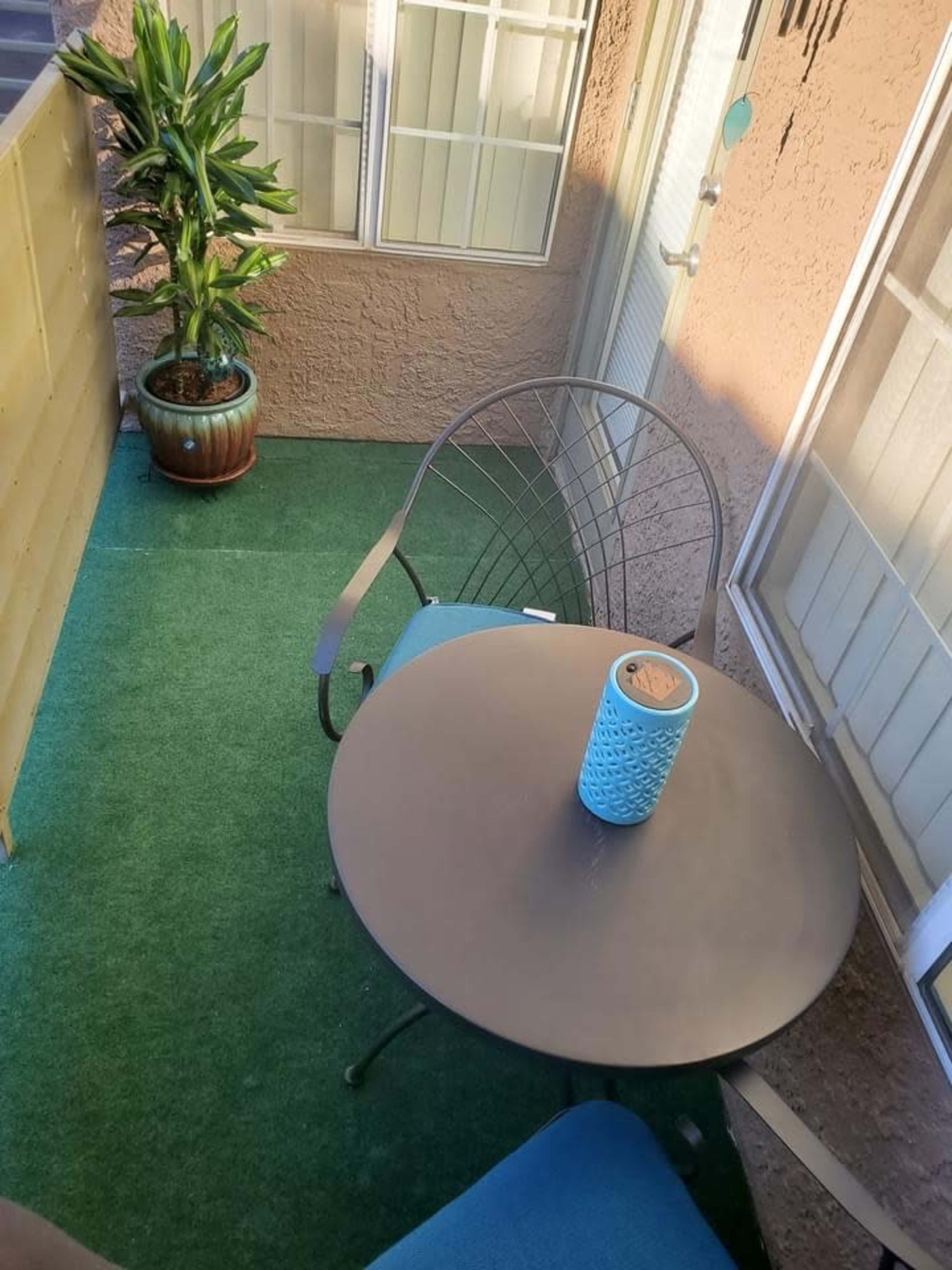 A small balcony features a round table with a blue decorative vase and one chair, surrounded by green artificial grass and a potted plant.