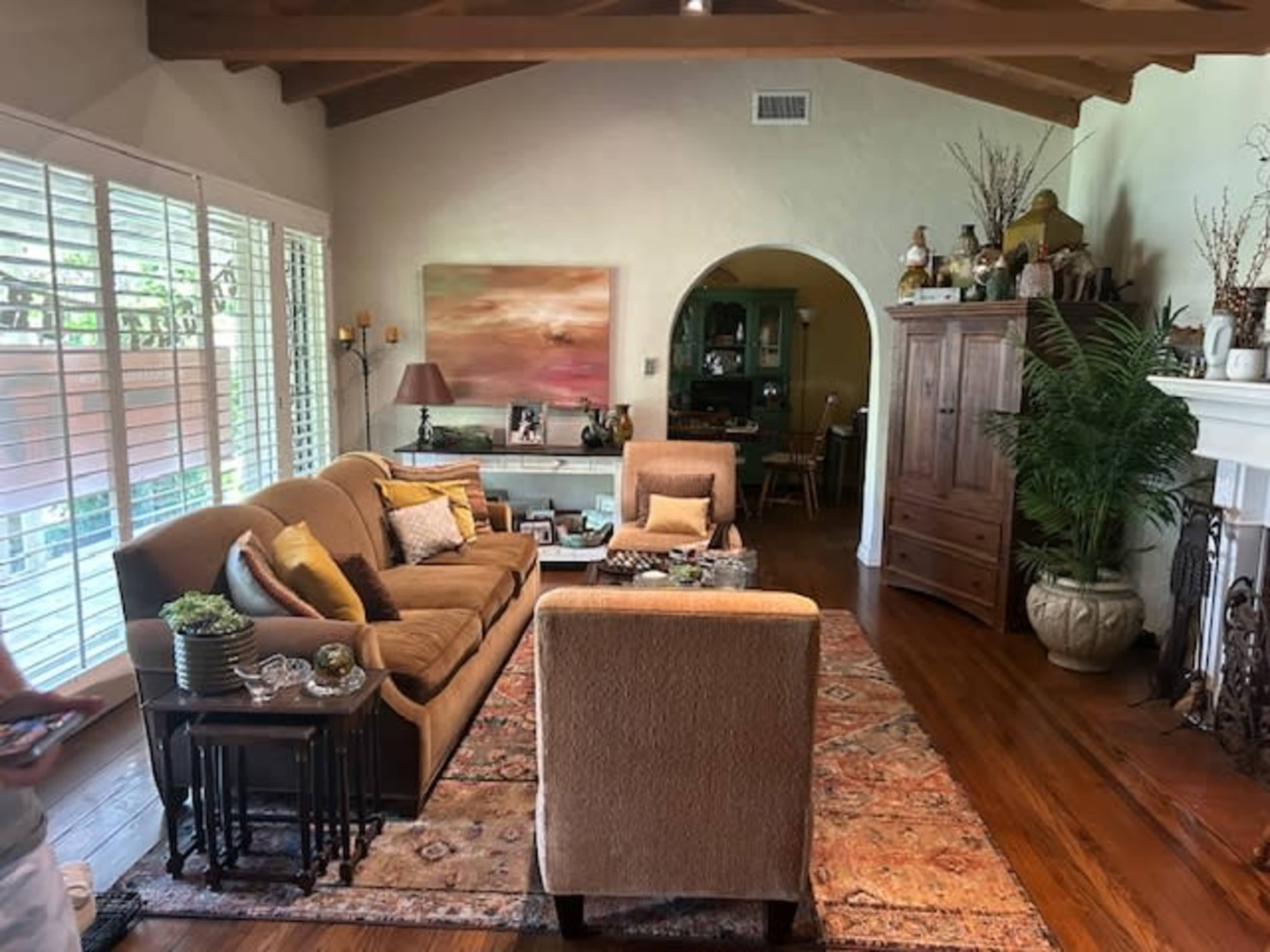 A cozy living room features a brown sofa, a matching armchair, and wooden furnishings, with natural light streaming through window shutters.