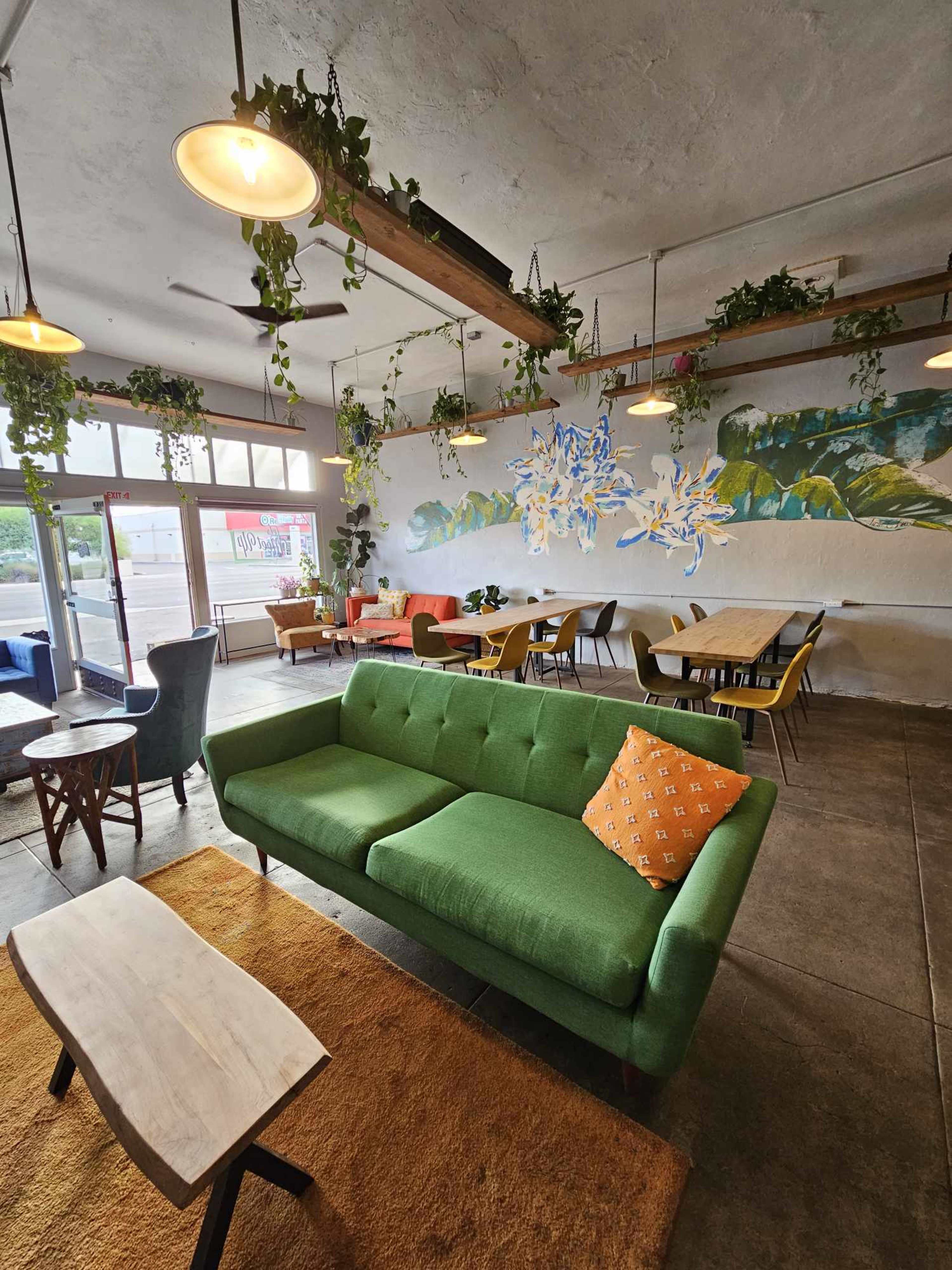A contemporary café interior with green sofas, wooden tables, and a mural of leaves on the wall, all complemented by hanging plants.
