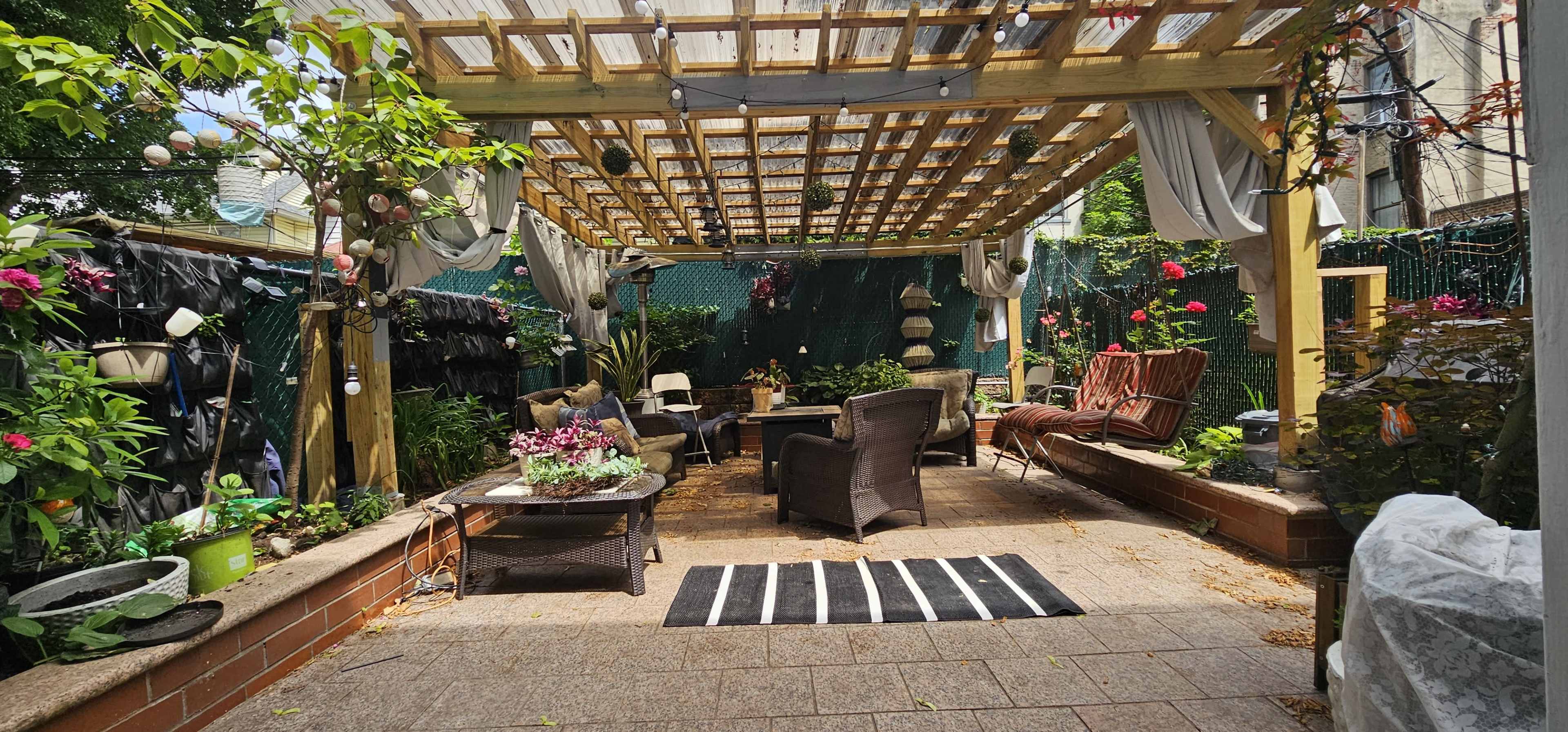 A landscaped patio area with a wooden trellis overhead, surrounded by potted plants and seating arrangements.