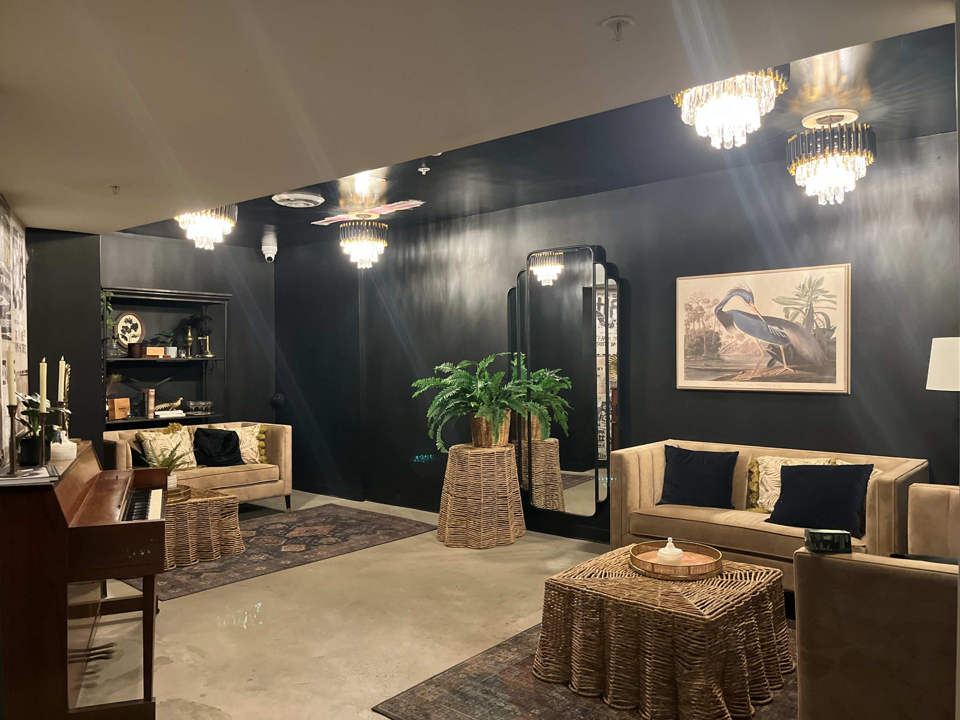 The image shows a stylish lounge area with two beige sofas, a mirrored wall, and a wooden piano, all illuminated by decorative chandeliers.