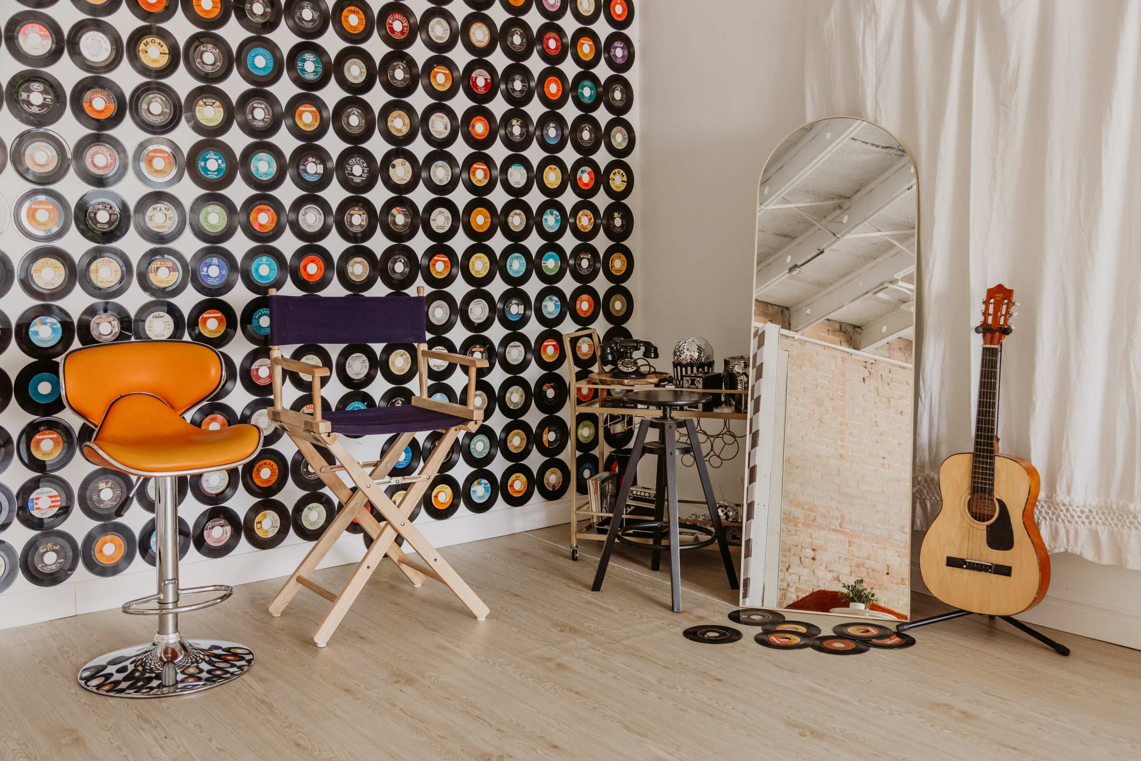 The image features a room with a wall decorated in colorful vinyl records, a wooden floor, a few stylish chairs, a tall mirror, and a guitar leaning against a chair.