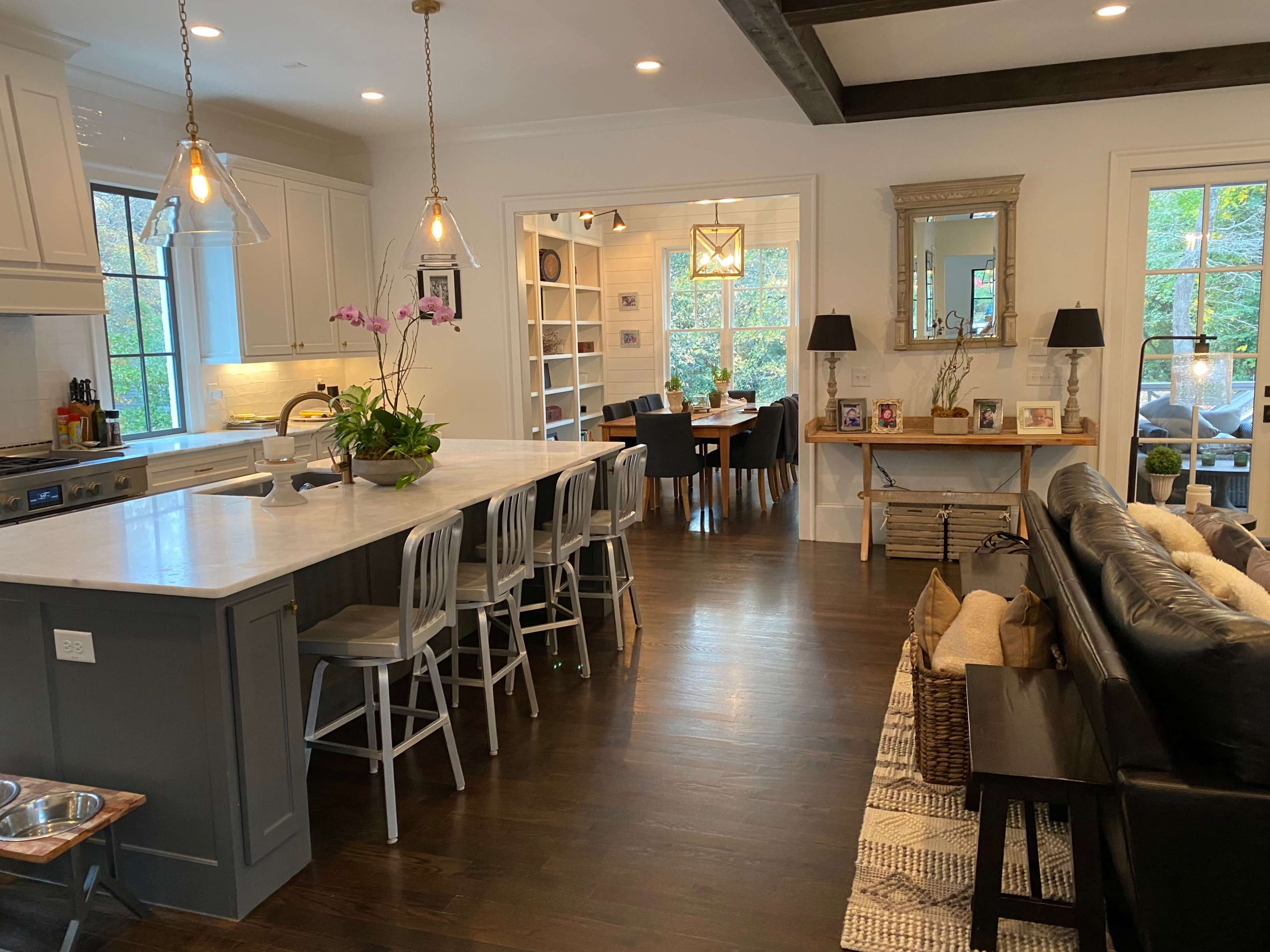 The image depicts a modern kitchen connected to a dining area, featuring a large island with bar stools, a dining table in the background, and natural light coming through windows.