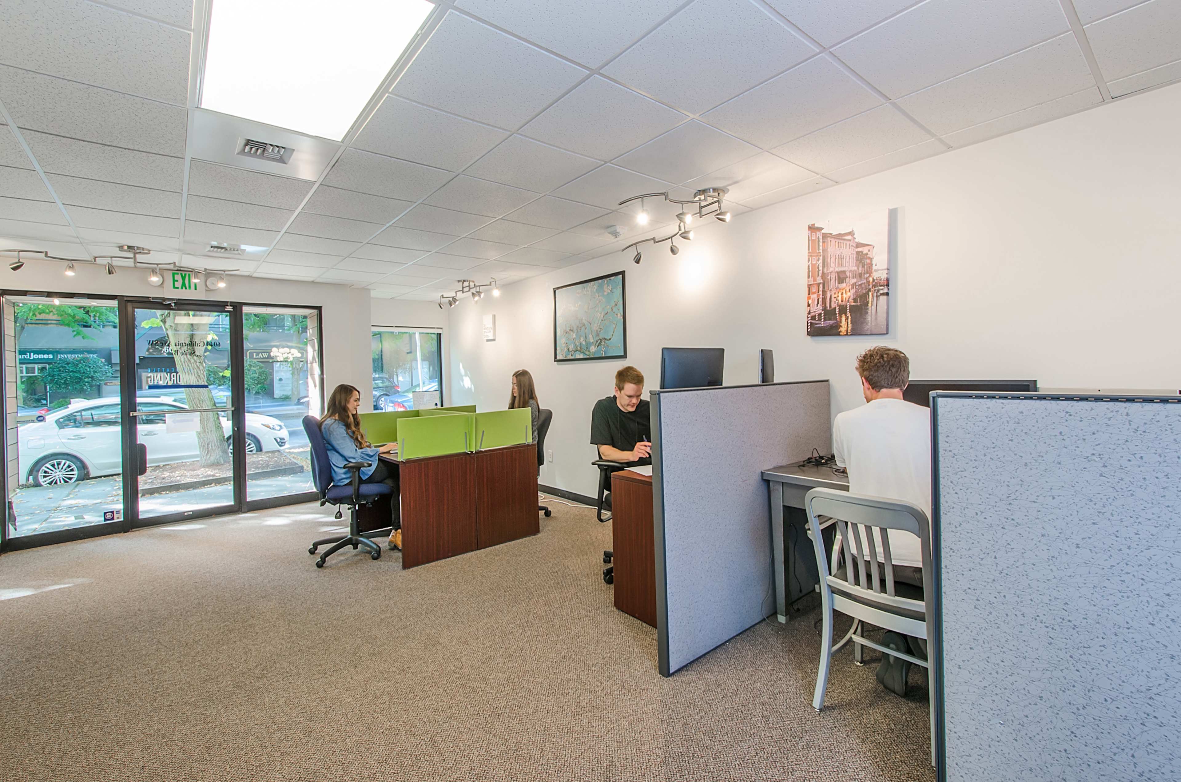 The image shows a modern office space with four workstations, two occupied by individuals at computers and two by people sitting in chairs, separated by low partitions.