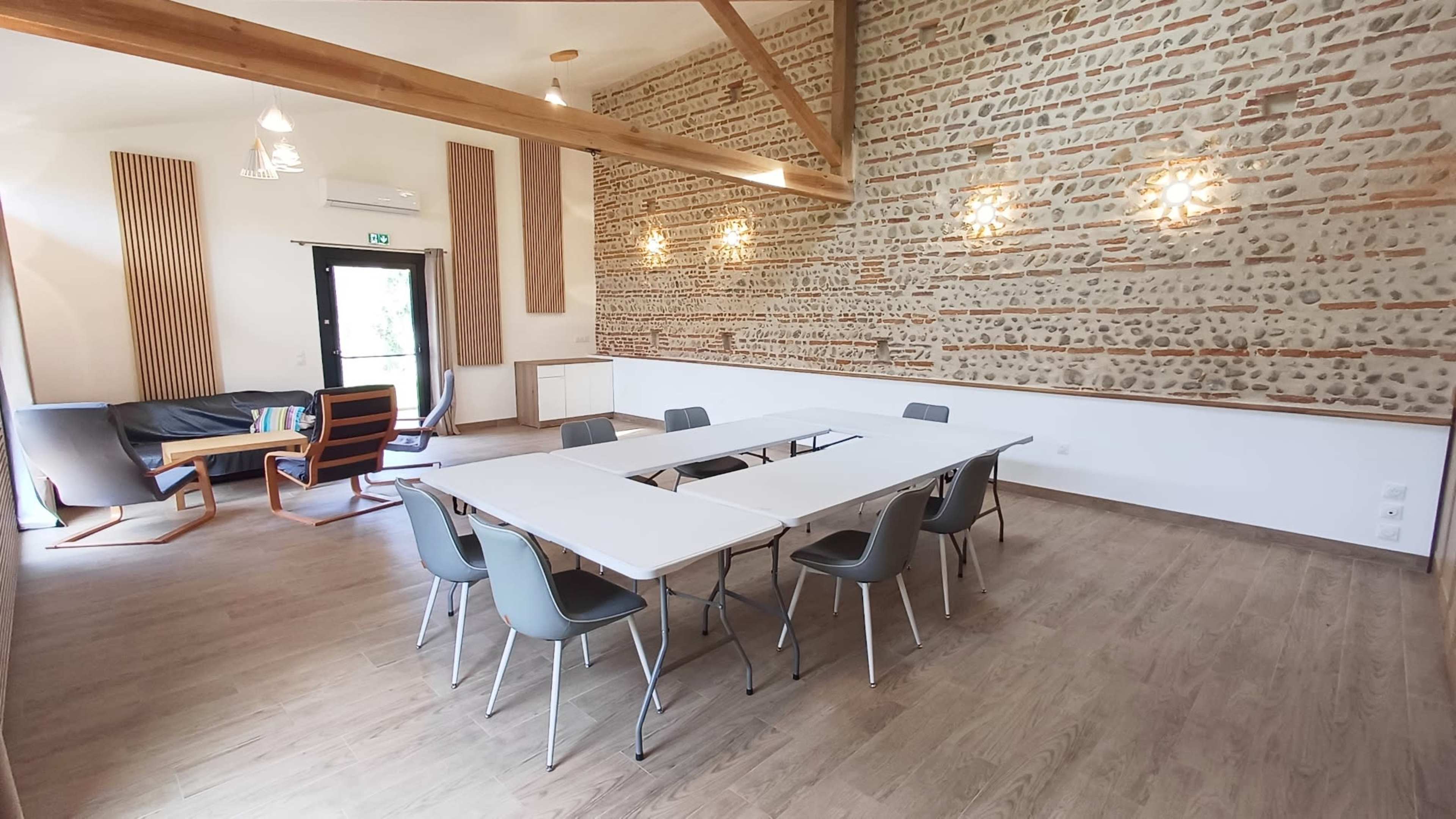 A bright meeting room features a white table surrounded by gray chairs, with a rustic stone wall and large windows.