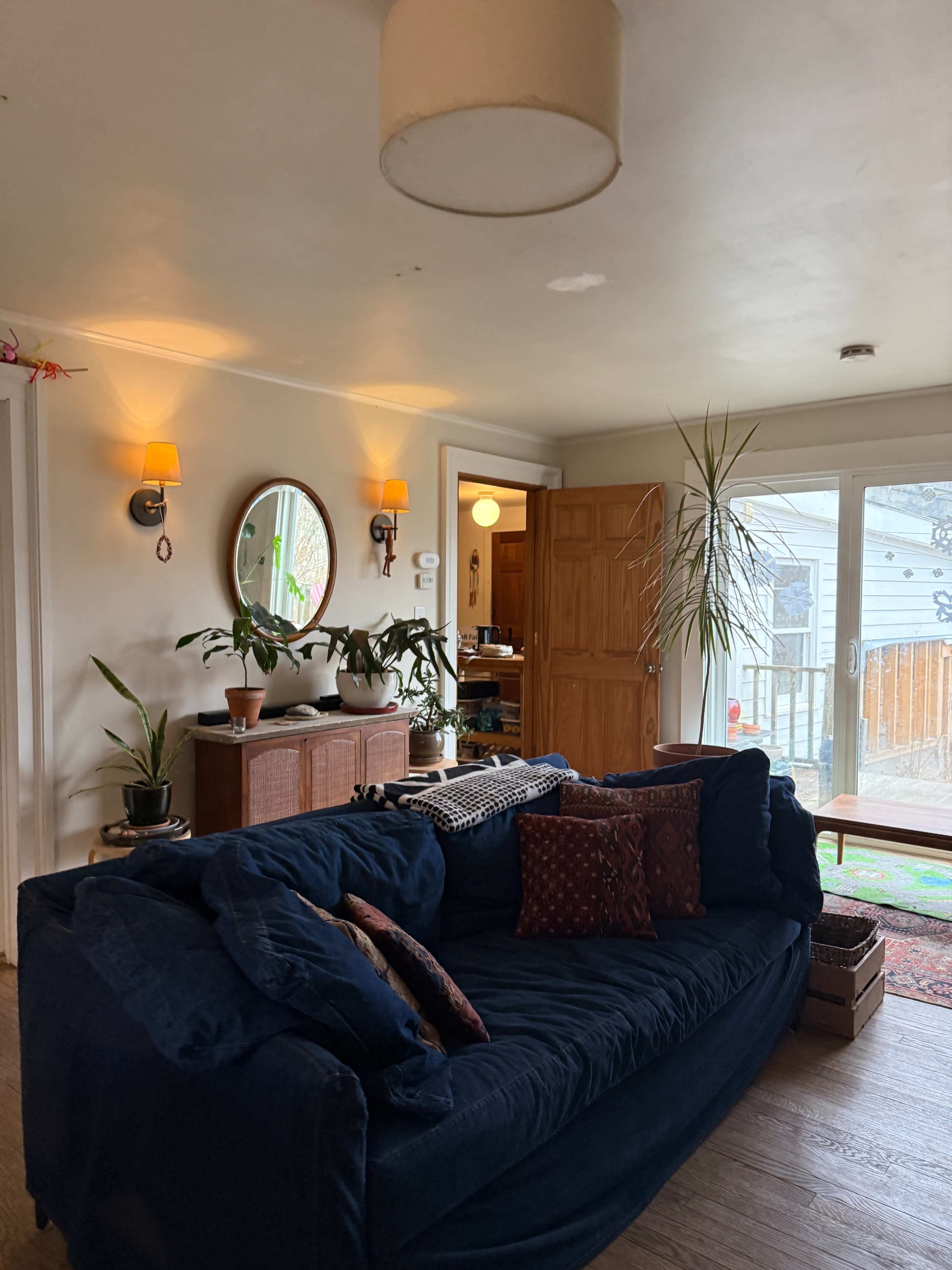 A blue couch is positioned in a well-lit living room adorned with plants, a round mirror, and wooden furniture.