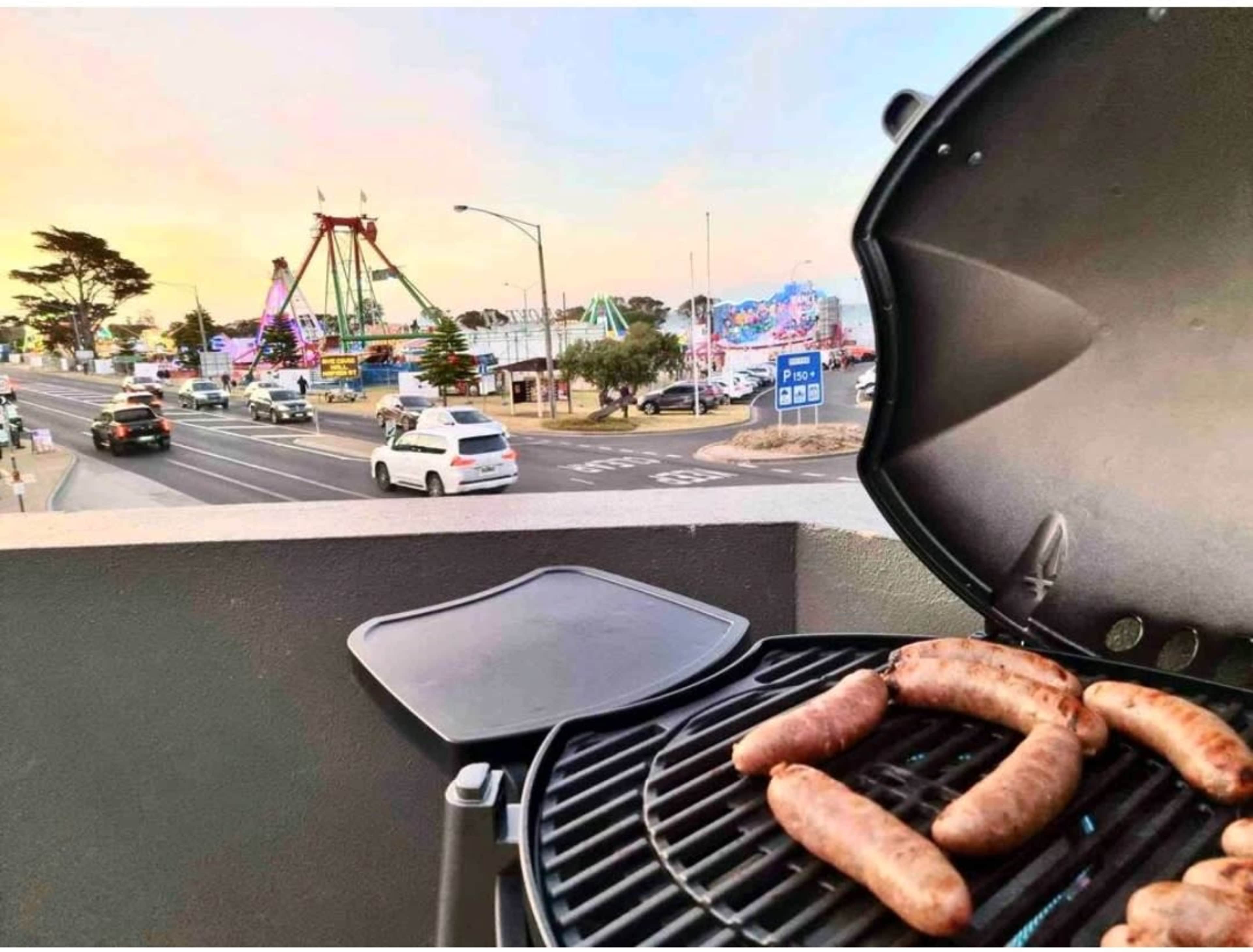 A barbecue grill with sausages is placed on a balcony overlooking a busy street and a fairground in the background.