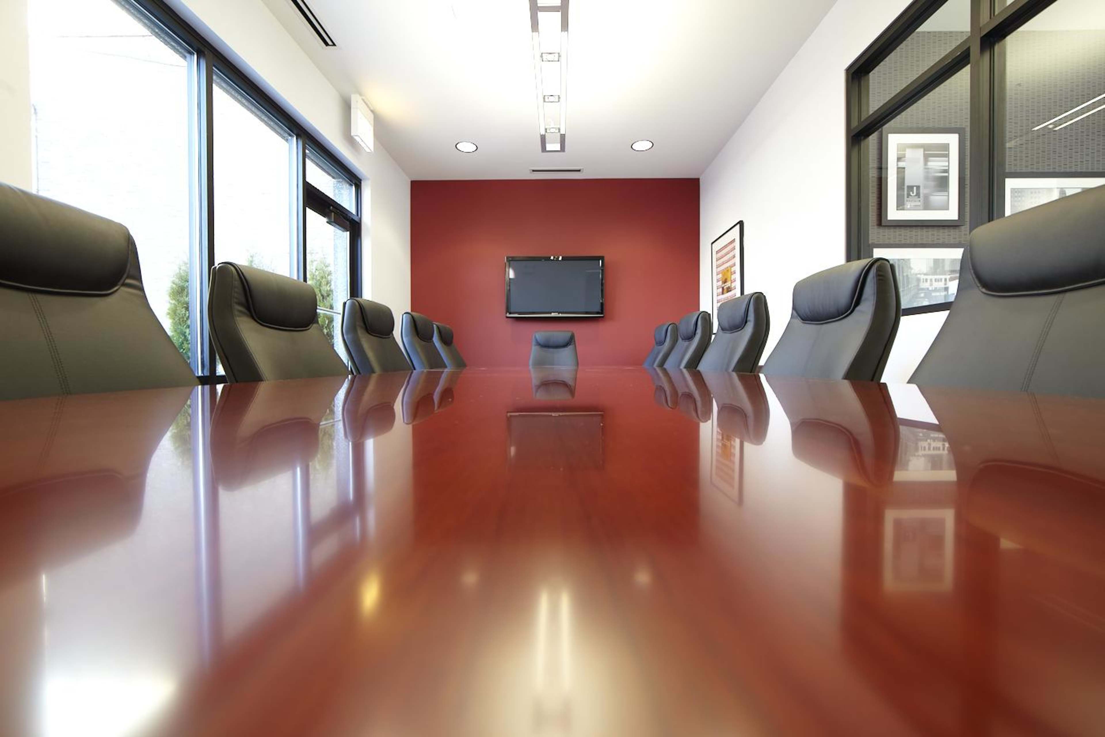 A long conference table with a polished surface is centrally positioned in a modern meeting room featuring dark leather chairs and a wall-mounted television.