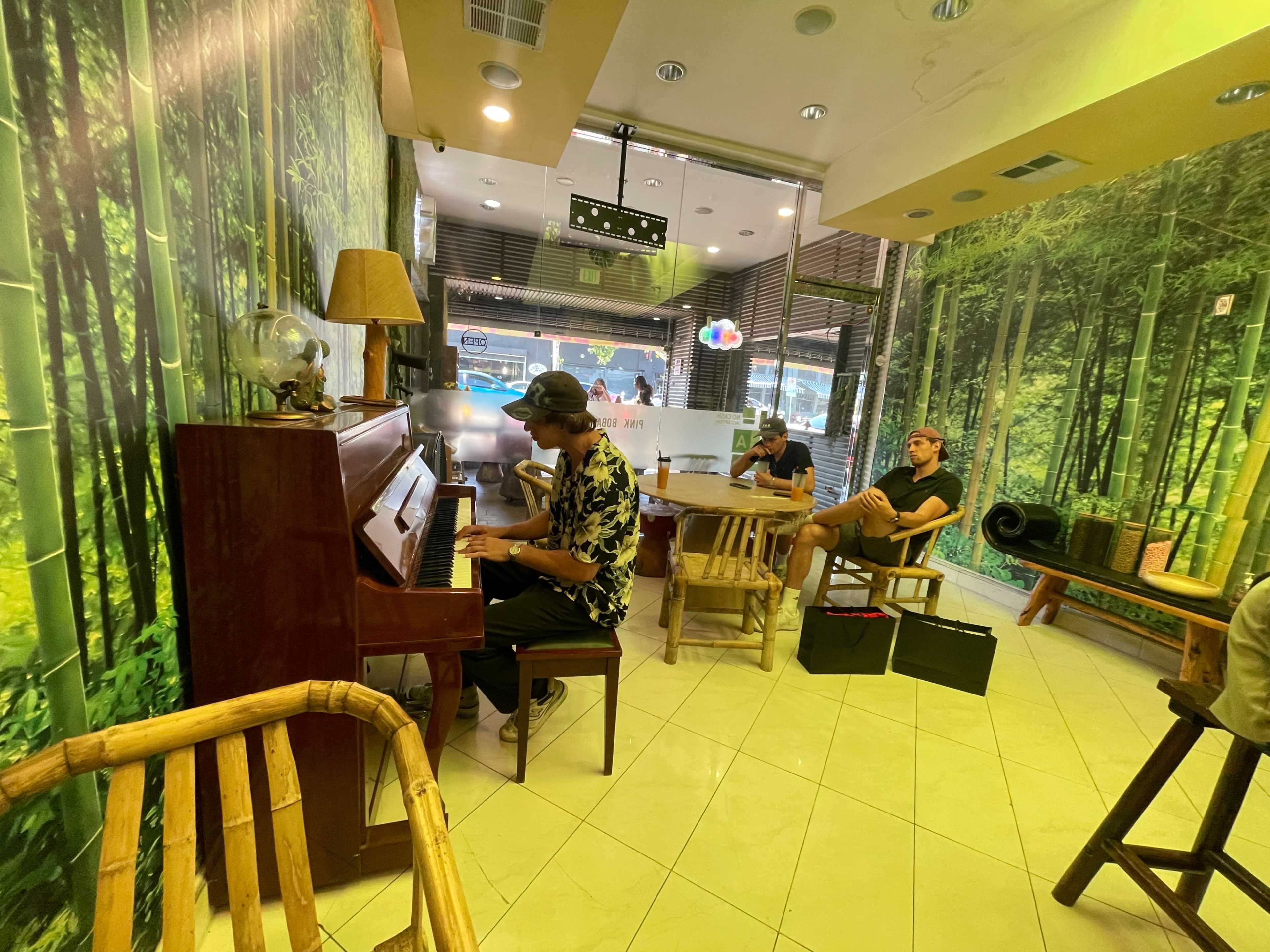 A musician plays a piano in a bamboo-themed cafe while two patrons sit nearby in wooden chairs.