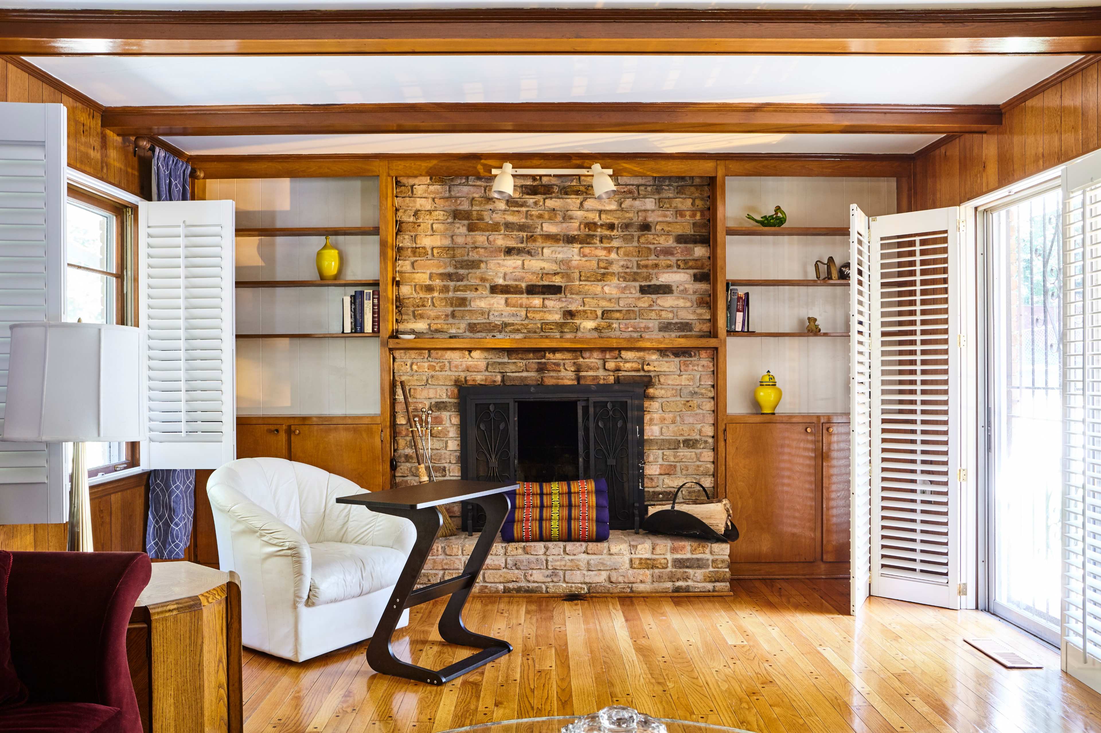 A living room featuring a brick fireplace, built-in shelves, a white armchair, and a wooden floor with large windows.