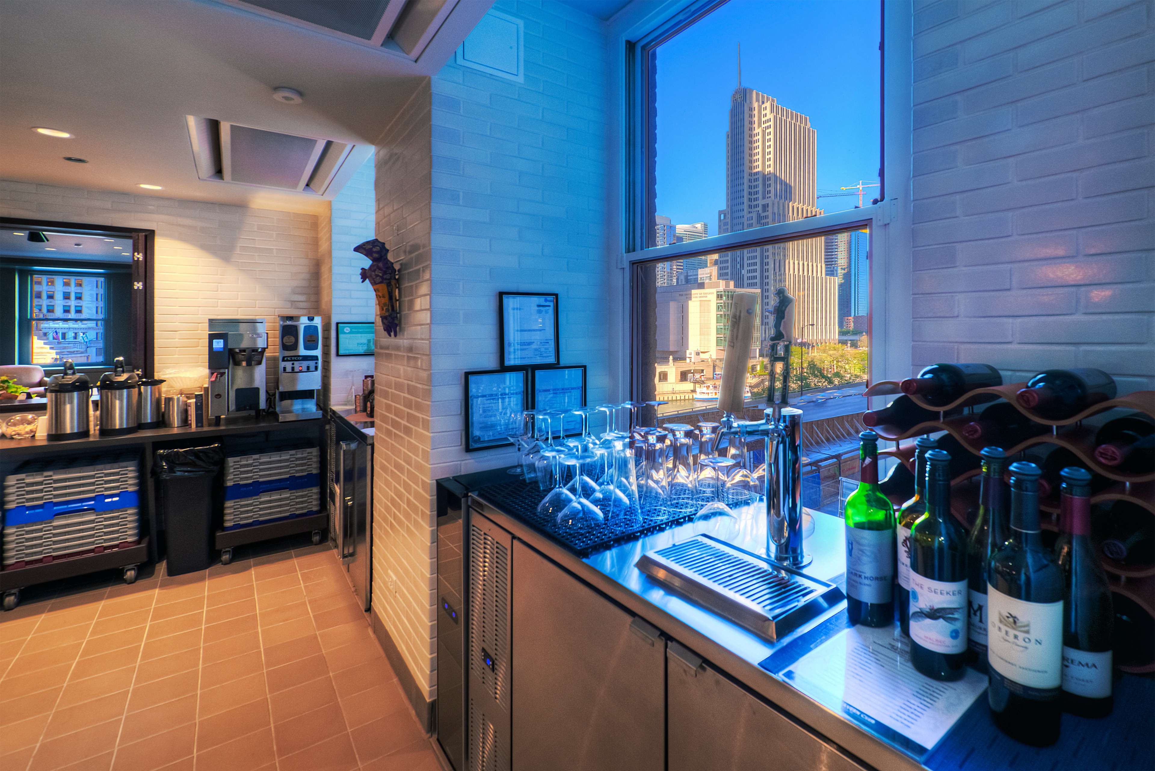 A modern kitchen area with a wine rack, glasses, and appliances, alongside a large window offering a view of a cityscape.