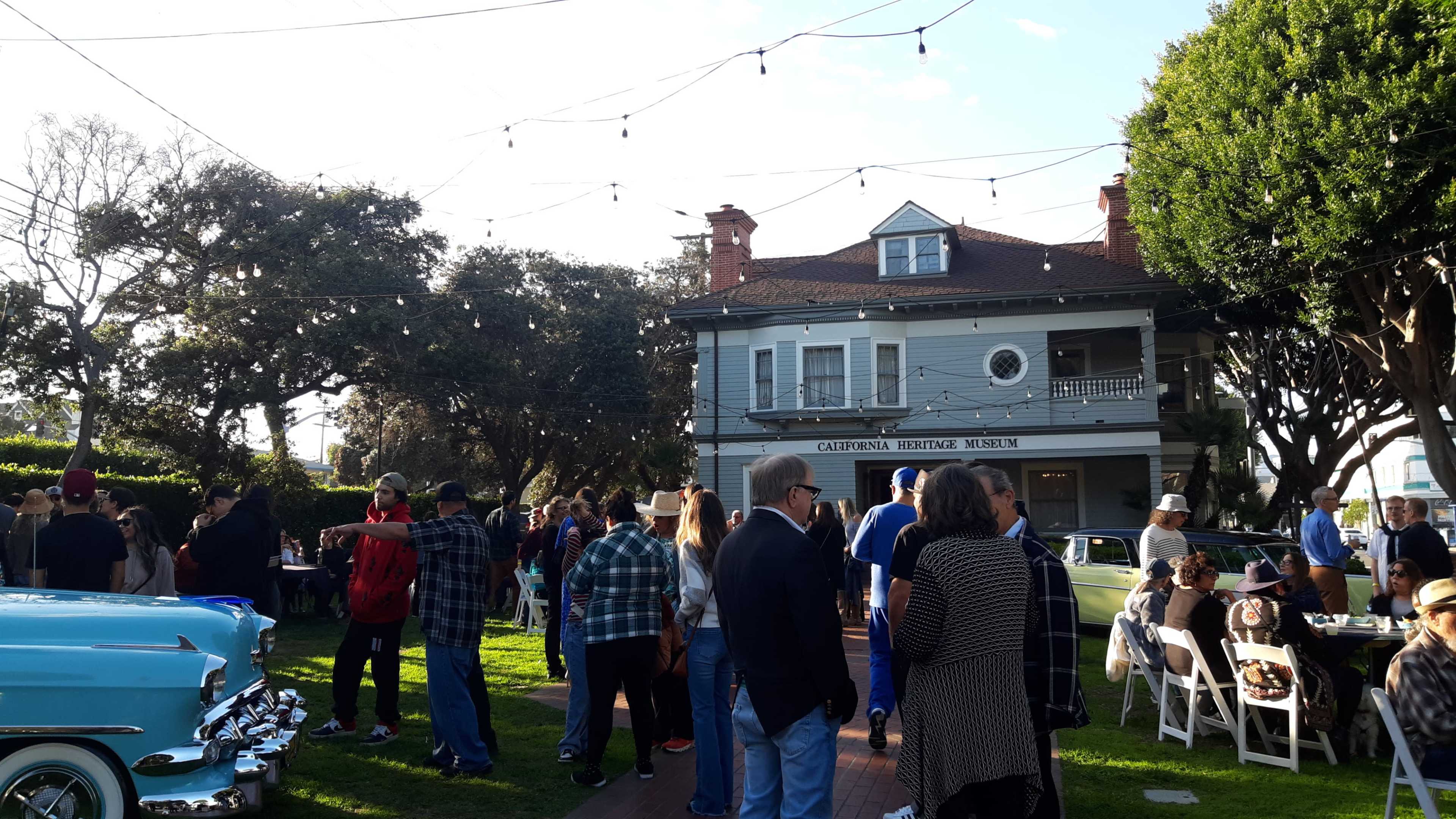 A crowd gathers at an outdoor event in front of the California Heritage Museum, surrounded by trees and vintage cars.