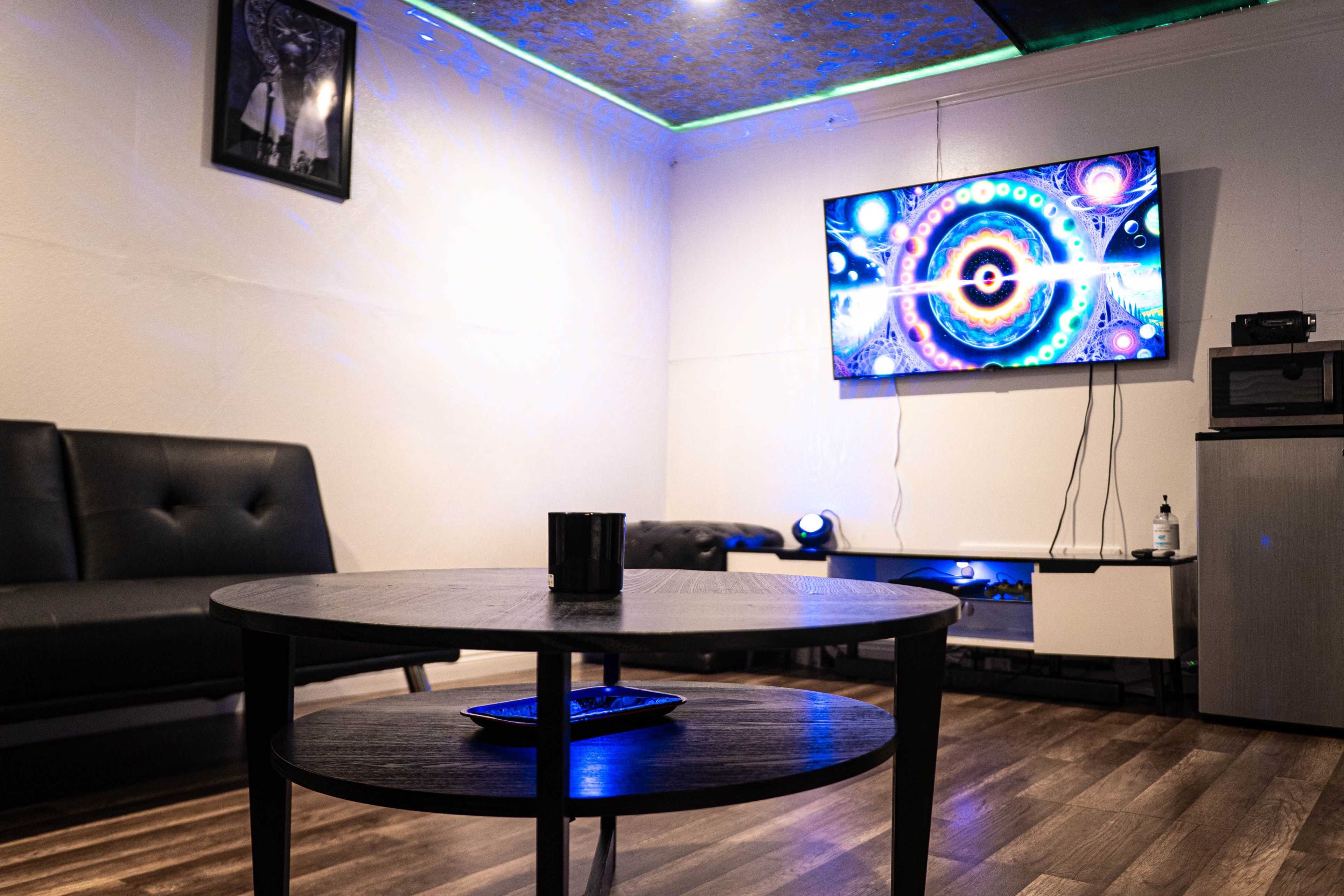 A modern, minimalist living room features a black couch, a round wooden coffee table with a remote, and a colorful abstract image displayed on a wall-mounted television.