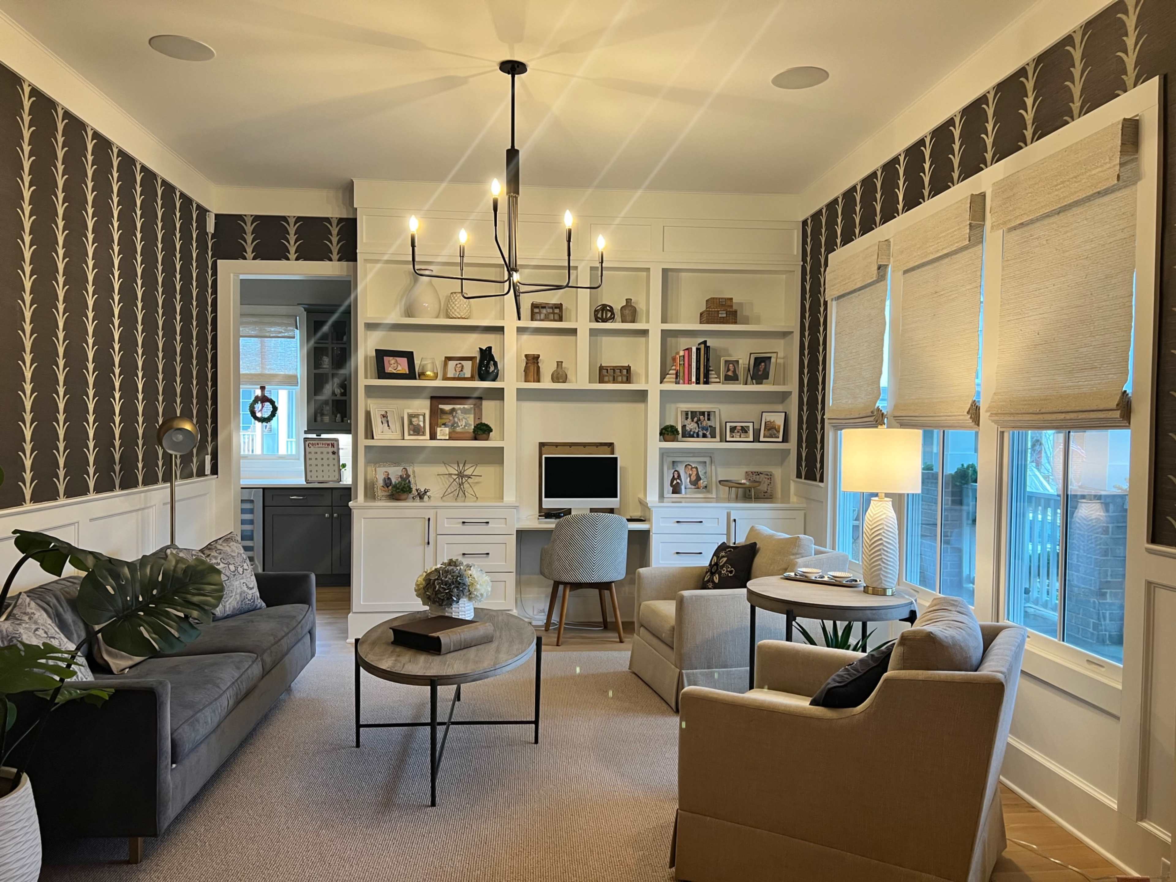 The image shows a stylish home office with a modern chandelier, built-in shelves filled with decor and books, and a seating area featuring two couches and a coffee table.
