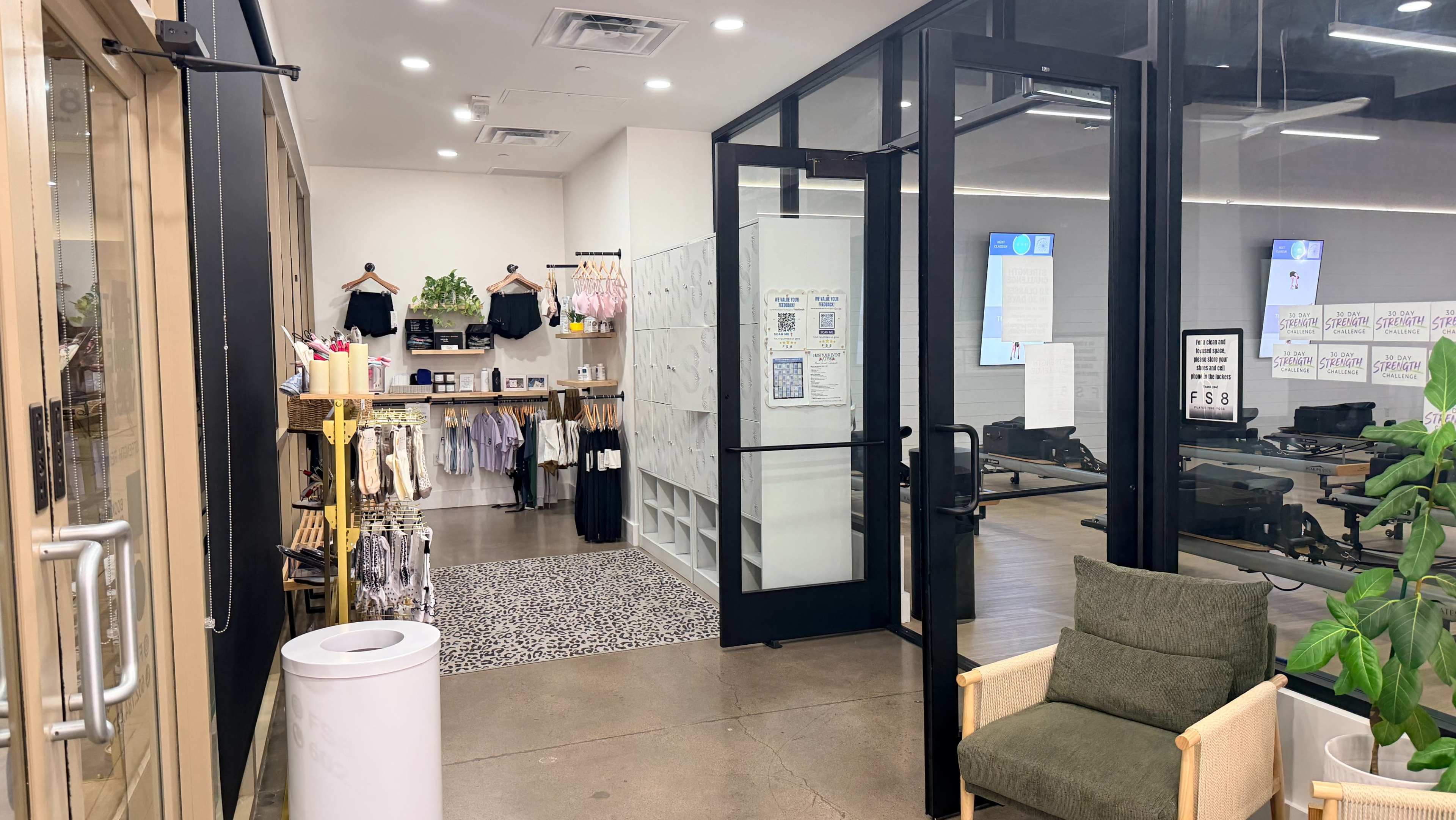 The image shows the interior of a fitness studio lobby, featuring a reception area with apparel displays, a waiting area with seating, and exercise equipment visible in the background.