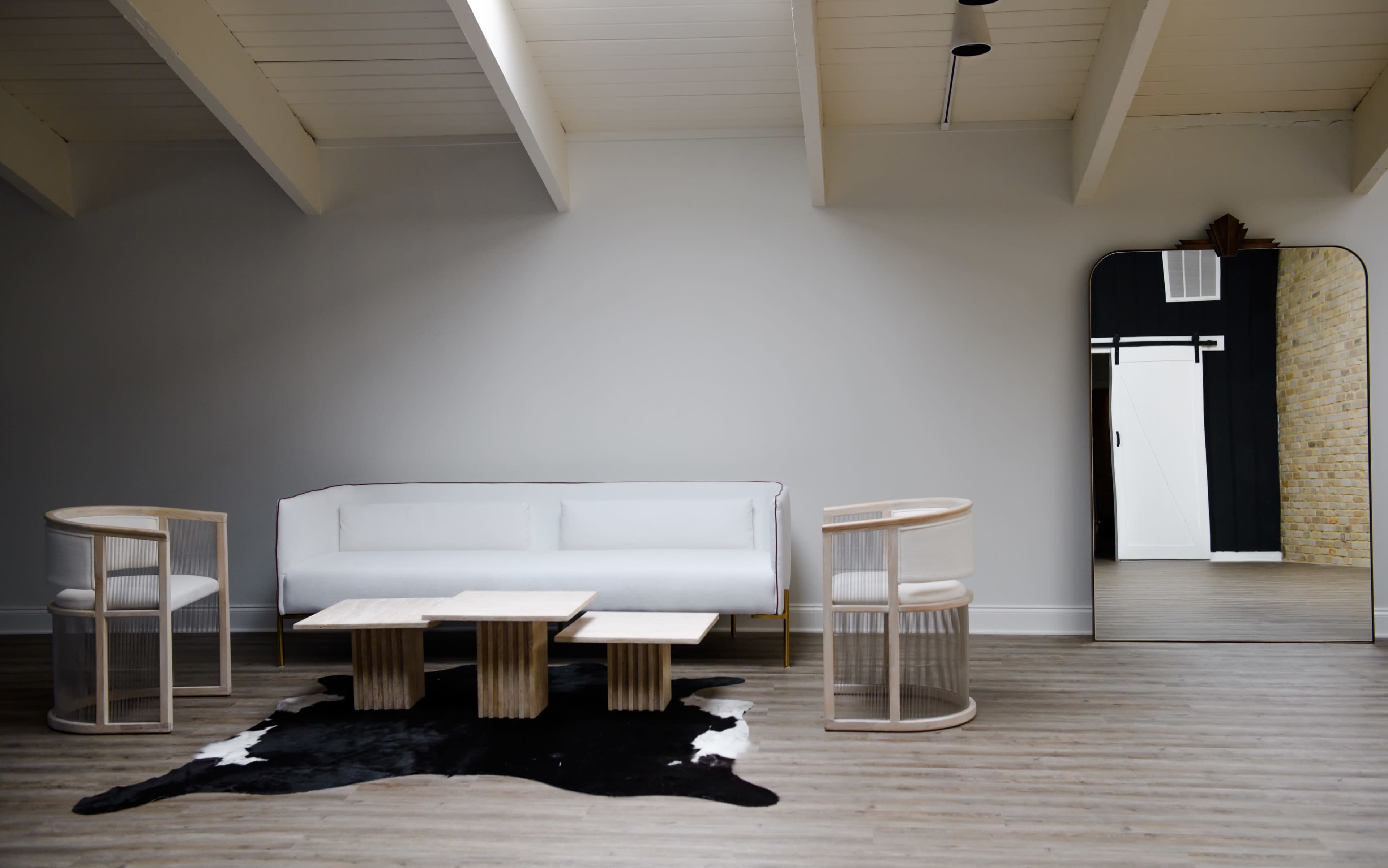 A minimalistic living space featuring a white sofa, two modern chairs, and a set of wooden coffee tables arranged on a patterned rug, with a large mirror reflecting a doorway in the background.