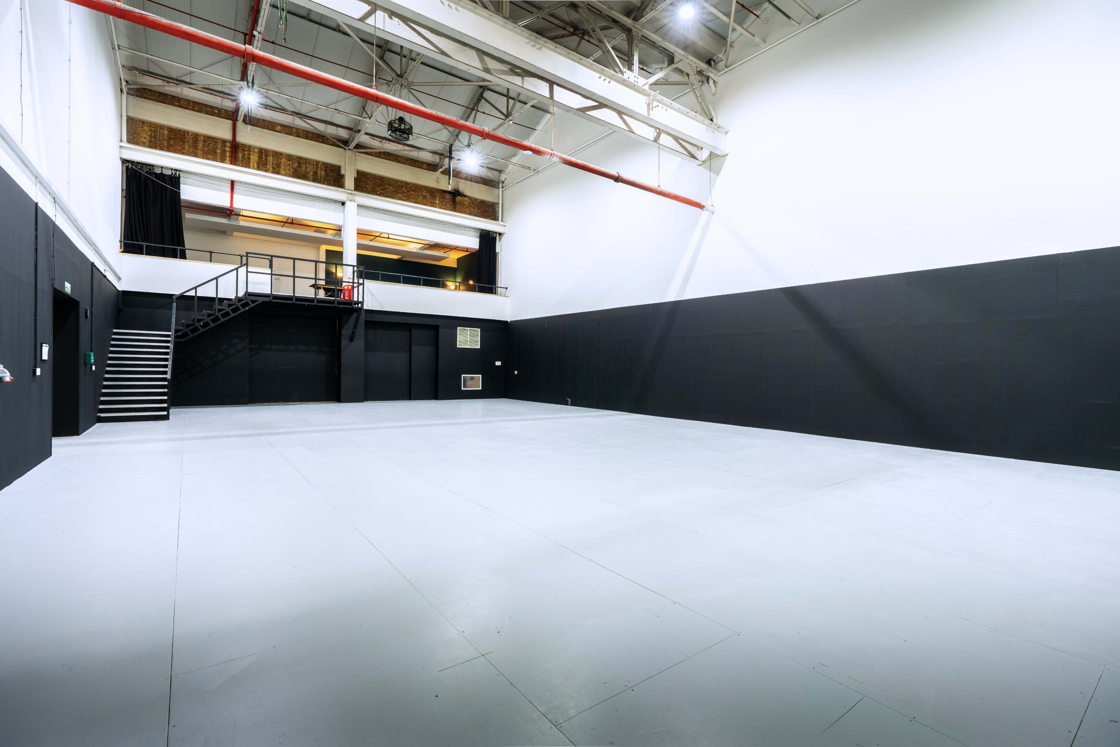 The image shows a large, empty studio space with high ceilings, black walls, and a staircase in the background.