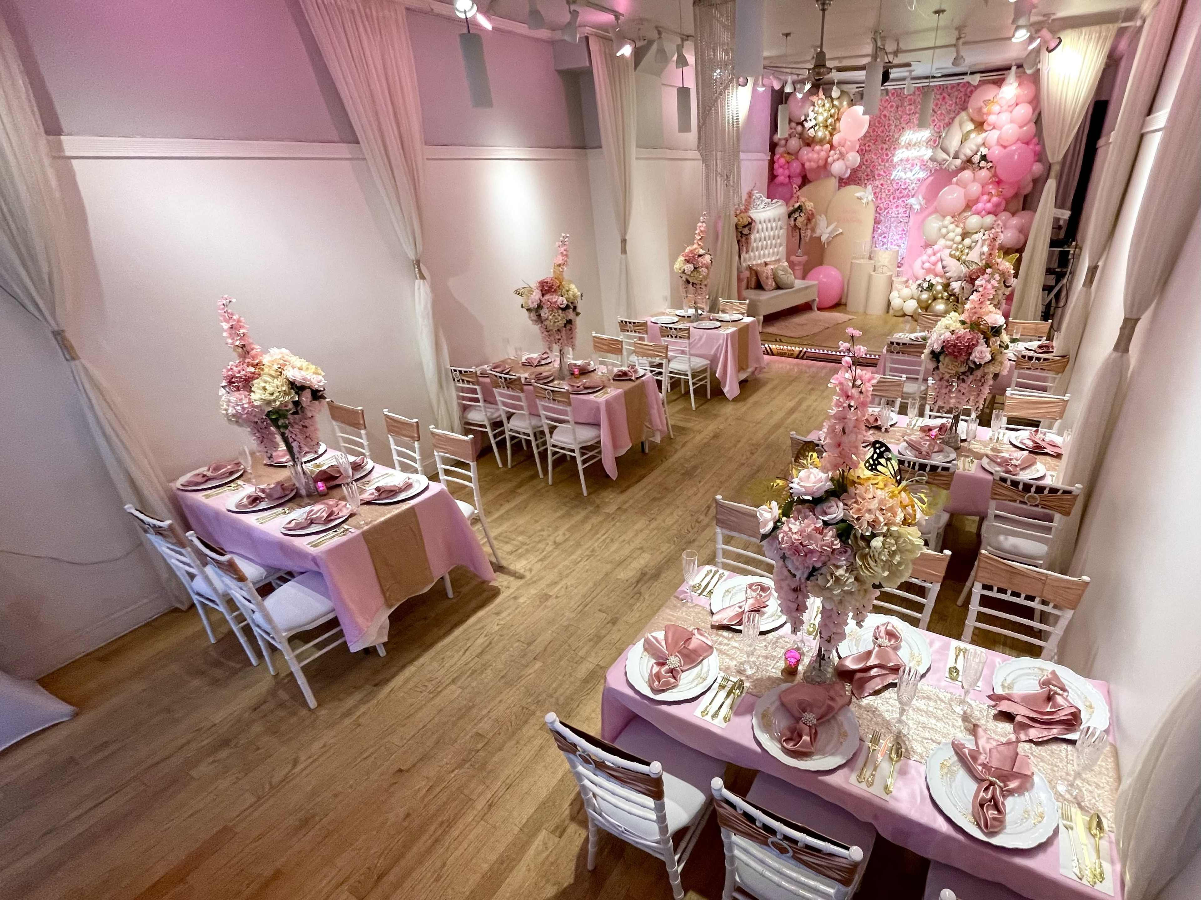 The image shows a light pink decorated event space with tables set for a celebration, featuring floral centerpieces and balloons in the background.