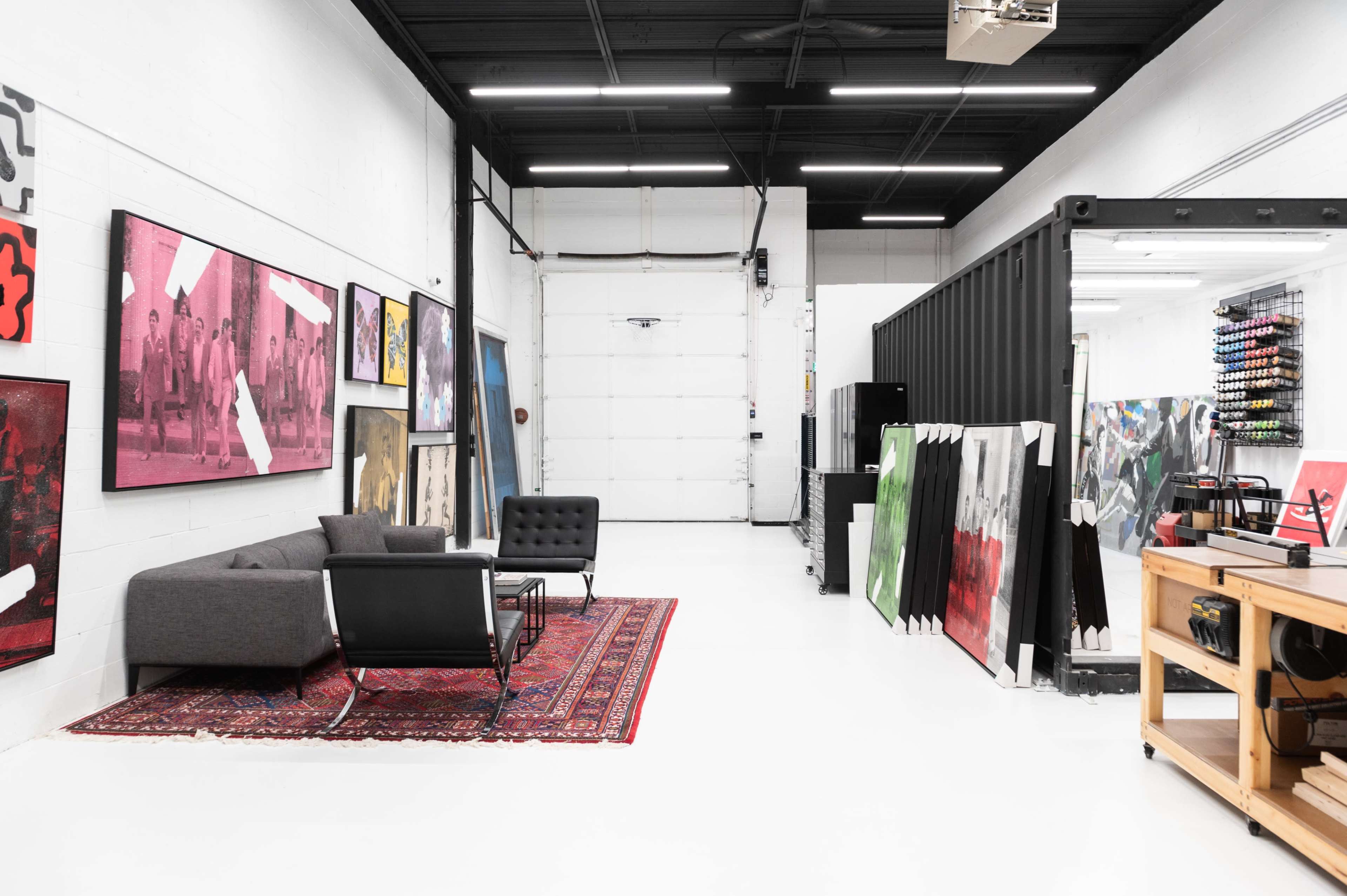An industrial art studio with a seating area featuring a gray sofa and black chairs, surrounded by colorful artworks and a large garage door.