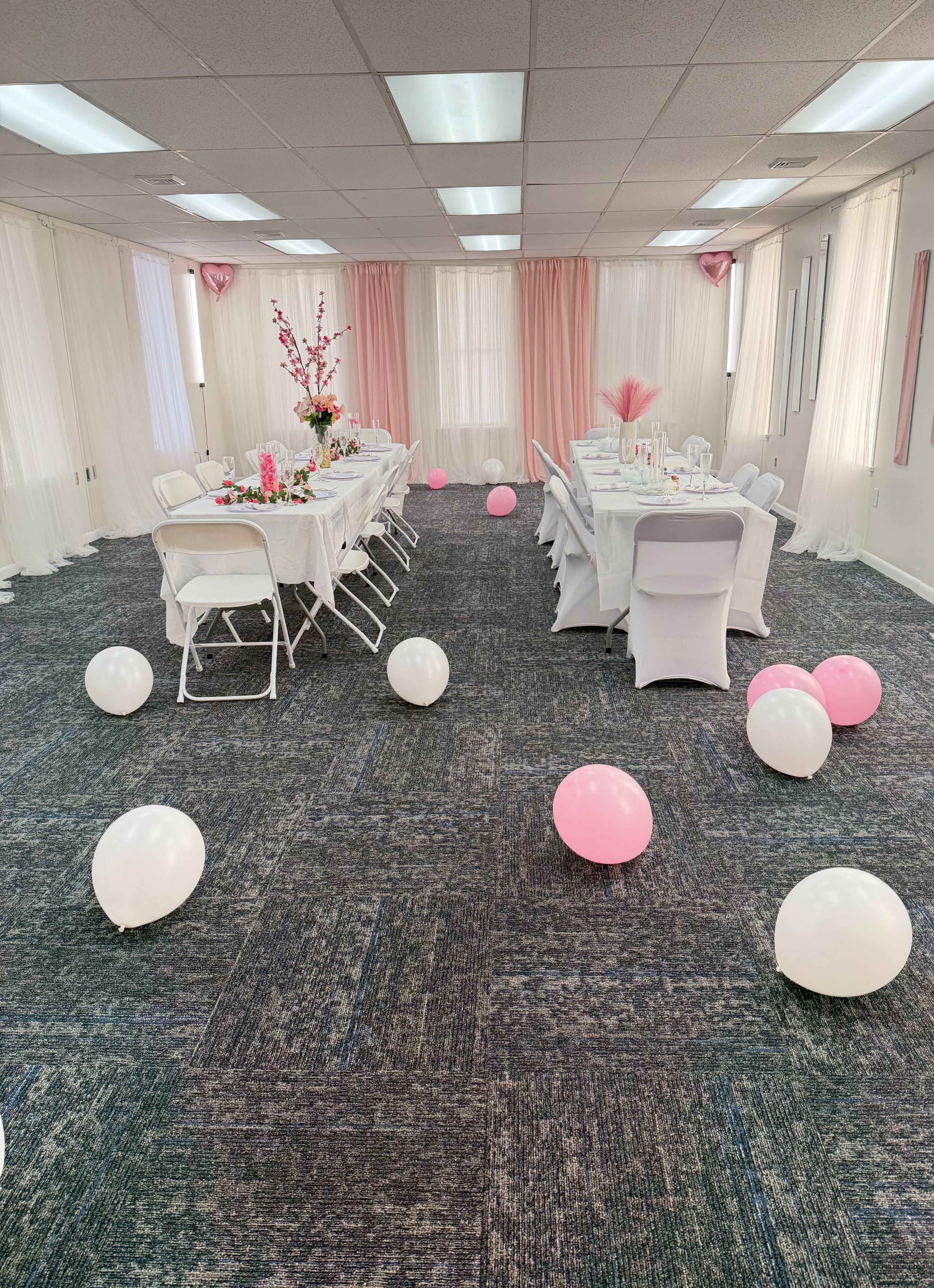 A party room is arranged with a long table set for guests, decorated with pink and white balloons scattered on the floor.