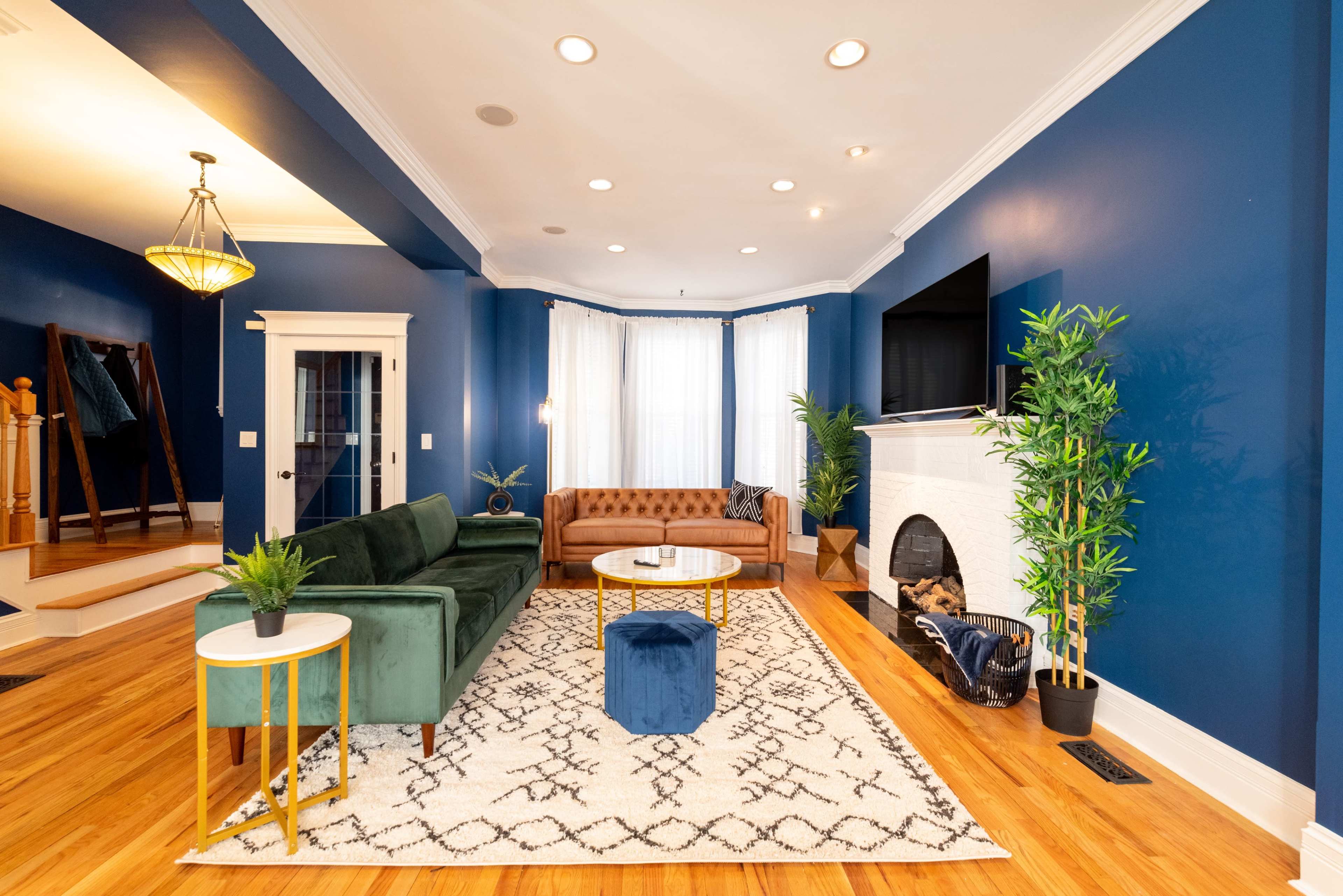 The room features a blue wall, a green sofa, a brown leather couch, a round coffee table, and a fireplace, with large windows allowing natural light to enter.