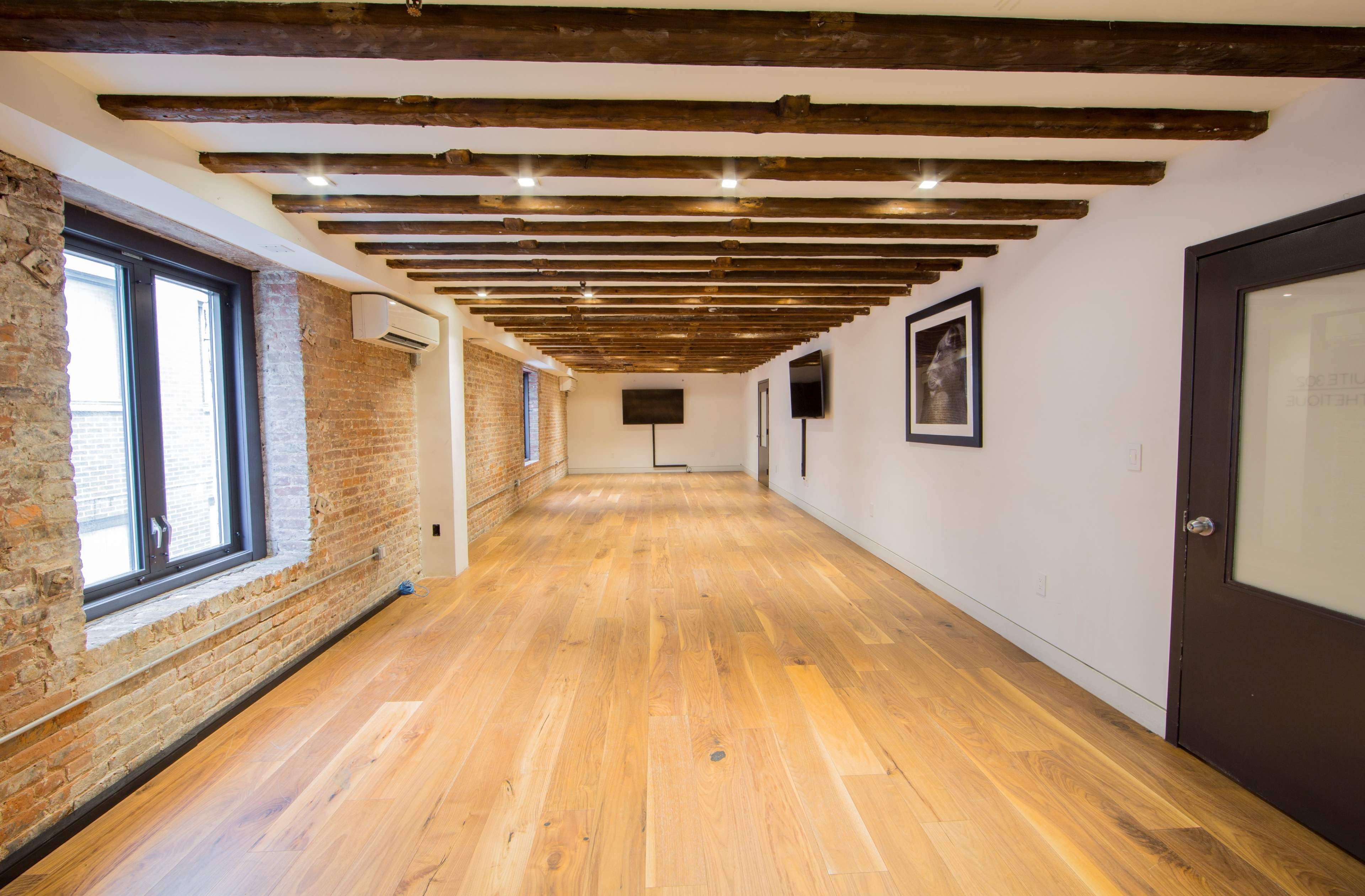 The image shows a spacious room with wooden beams on the ceiling, large windows, and hardwood flooring.