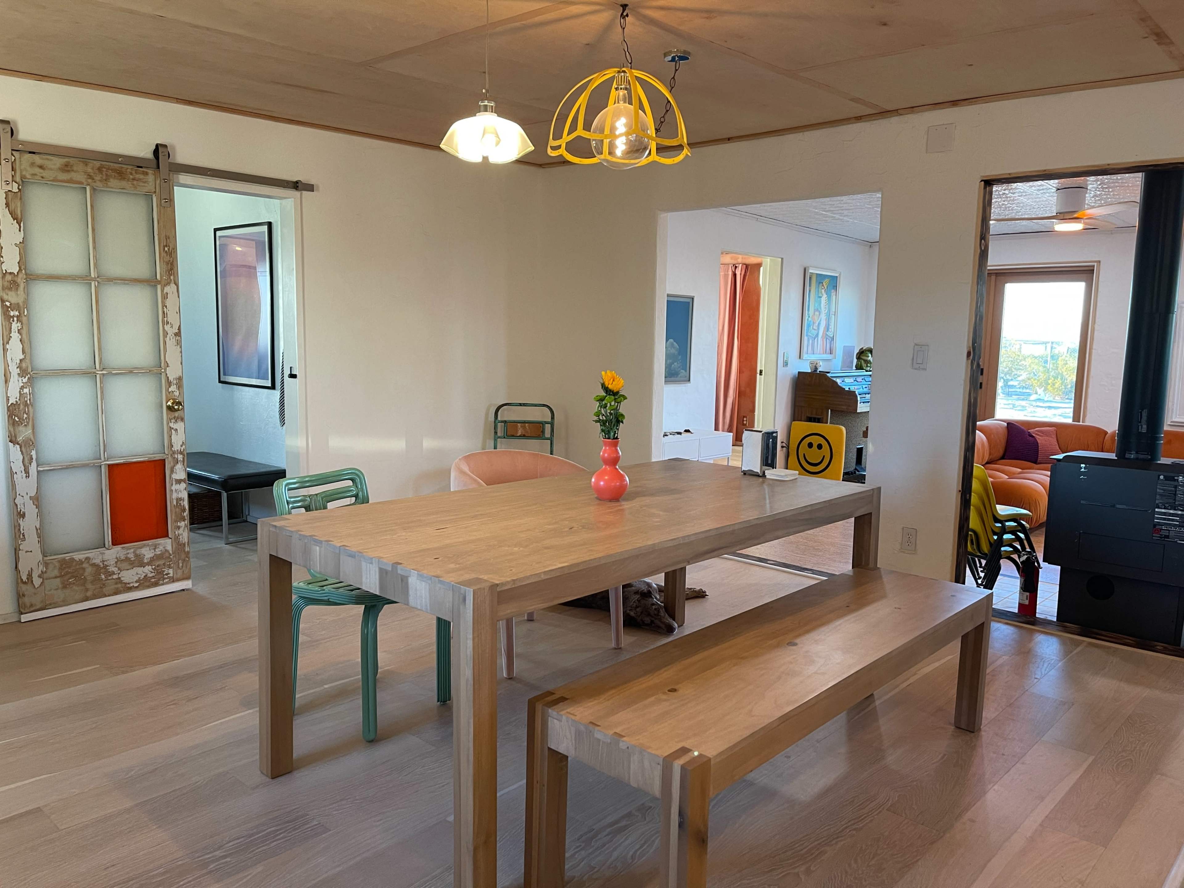 A wooden dining table with a vase of flowers sits between a bench and a chair in a brightly lit room featuring a mix of colorful furnishings and a sliding door.