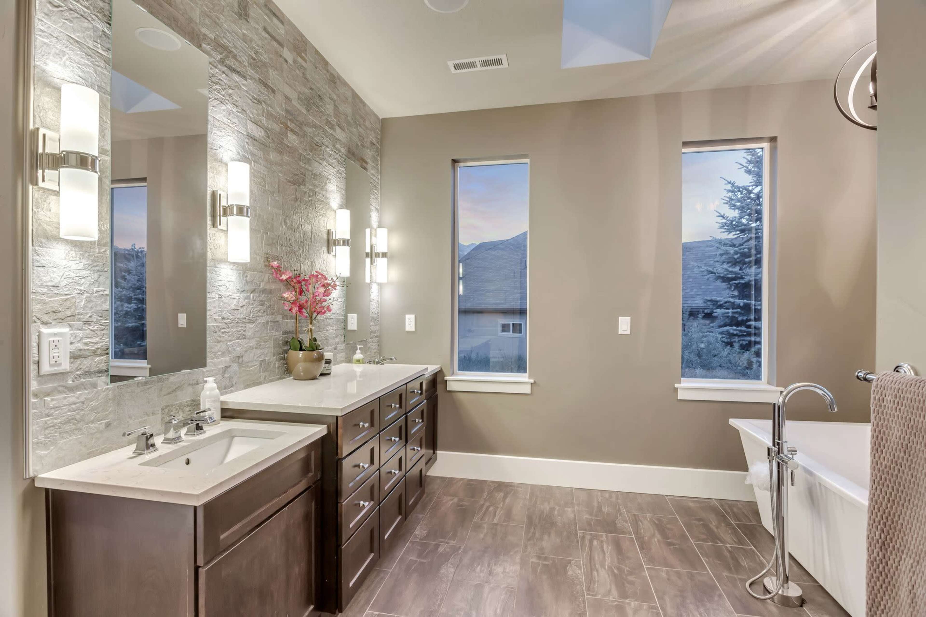 A modern bathroom featuring a double vanity with two mirrors, stone accent walls, and a freestanding bathtub next to a window.