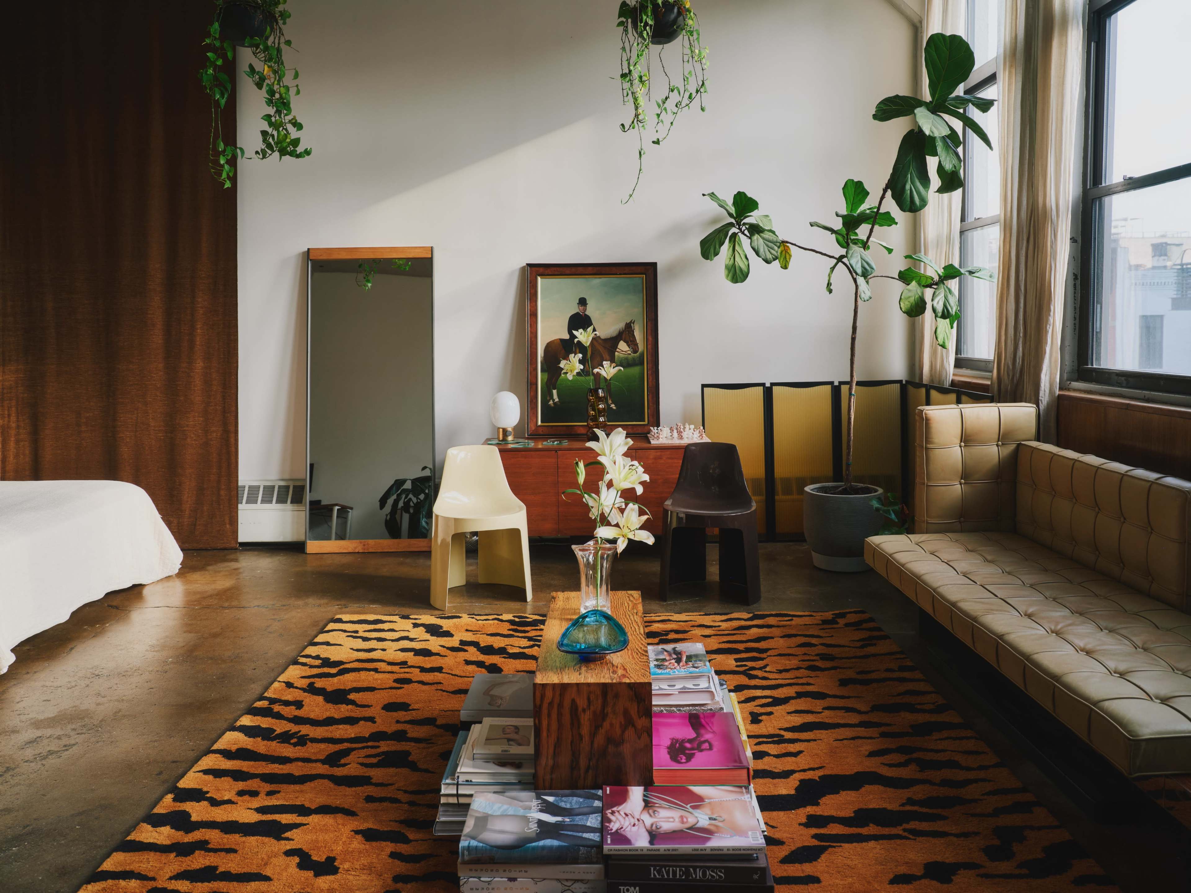 The scene shows a modern living space with a leather sofa, a coffee table stacked with magazines, a large mirror, and several plants hanging from the ceiling.