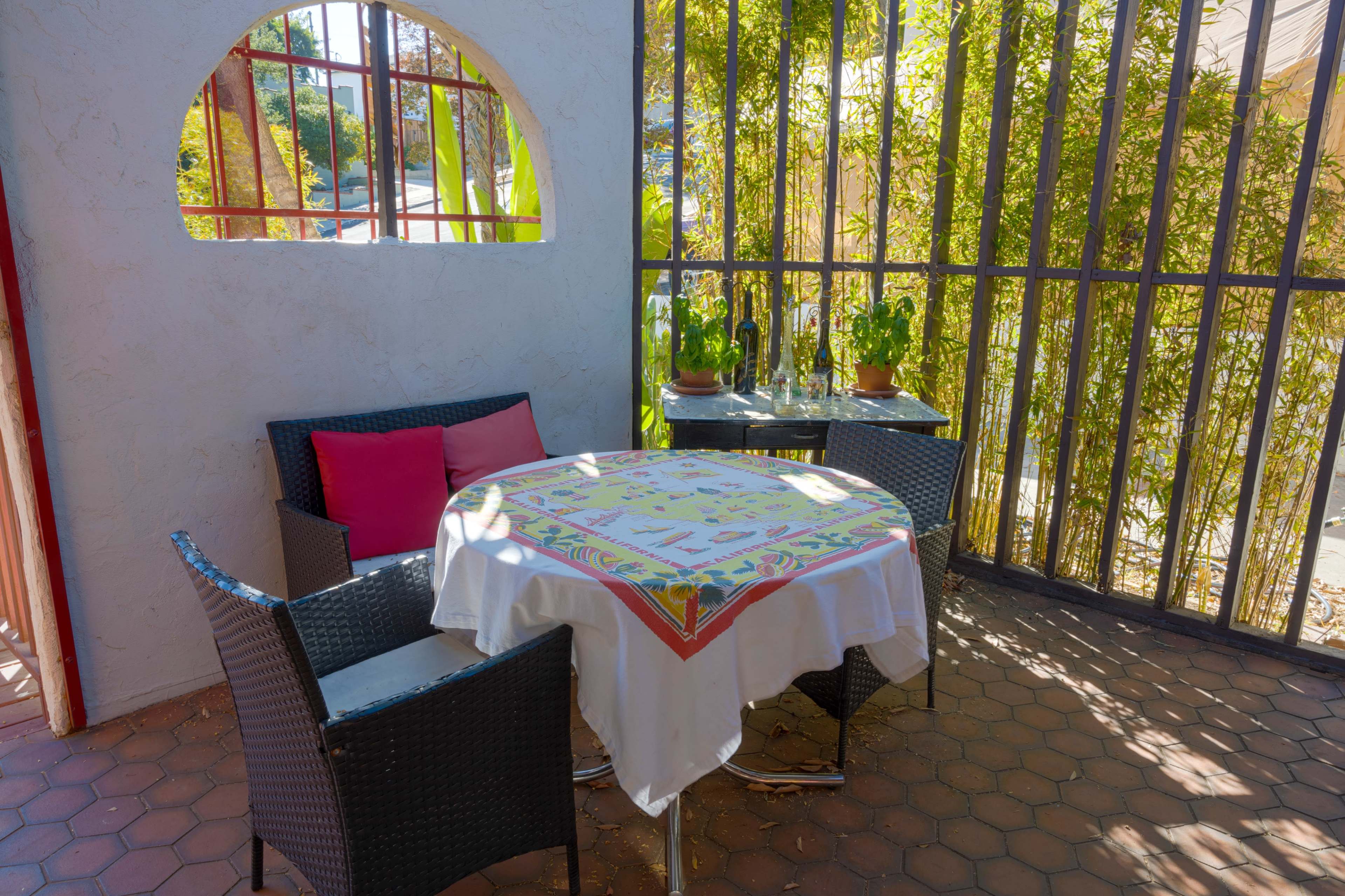 A small outdoor seating area features a round table covered with a tablecloth and surrounded by two wicker chairs, with potted plants visible in the background.
