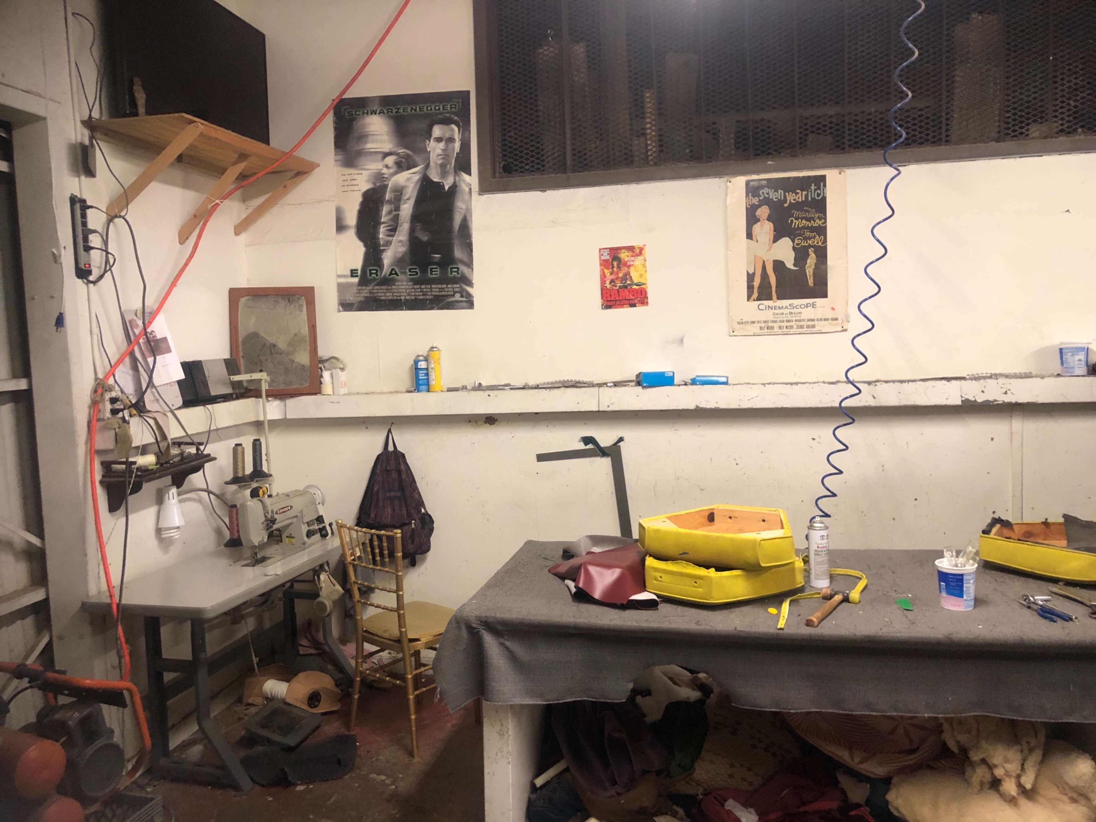 The image shows a cluttered workshop with a sewing machine, a table covered in materials, and various posters on the walls.