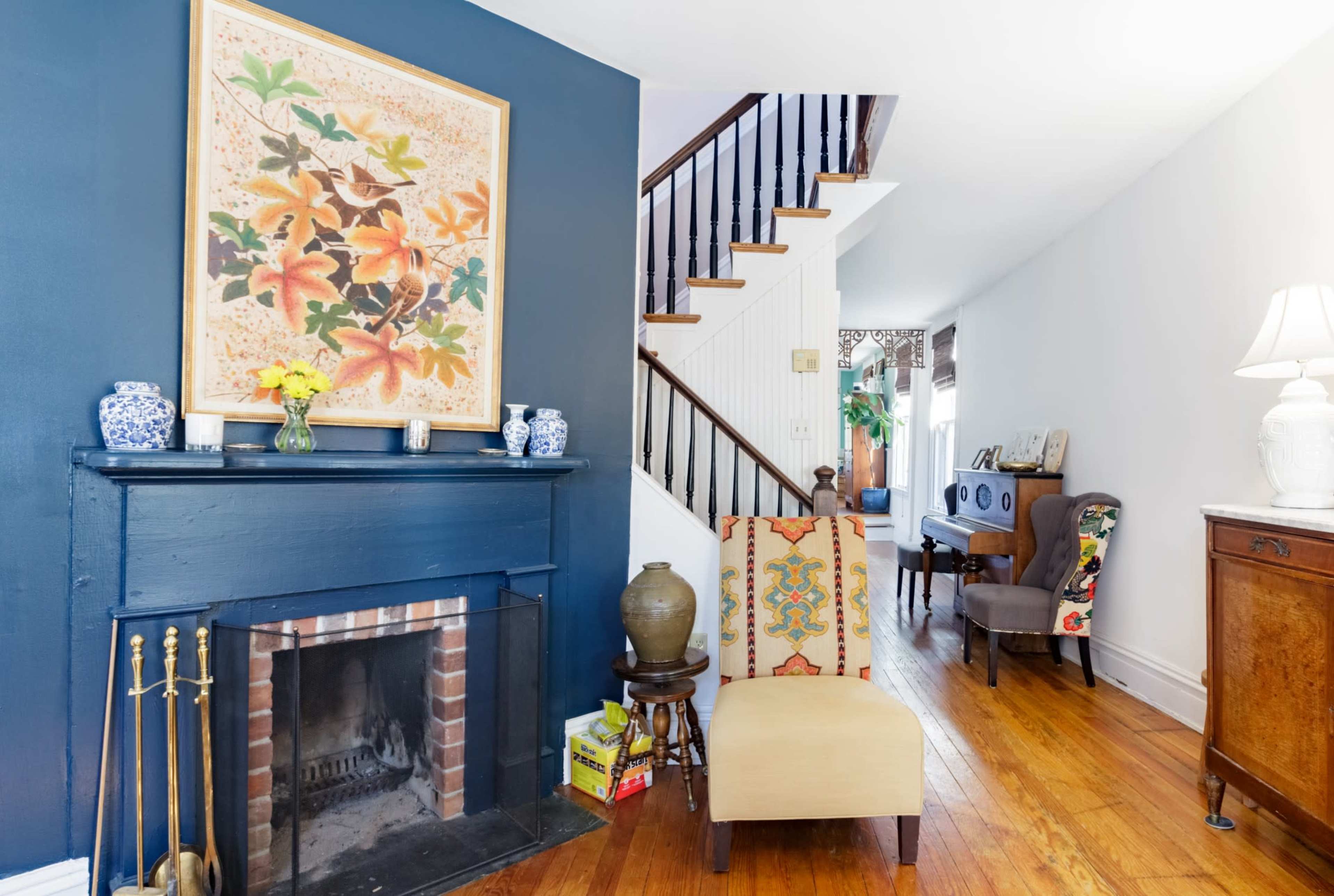 A living room with a fireplace, a colorful flower painting on the blue wall, a staircase in the background, and an arrangement of furniture including a chair and decorative items.