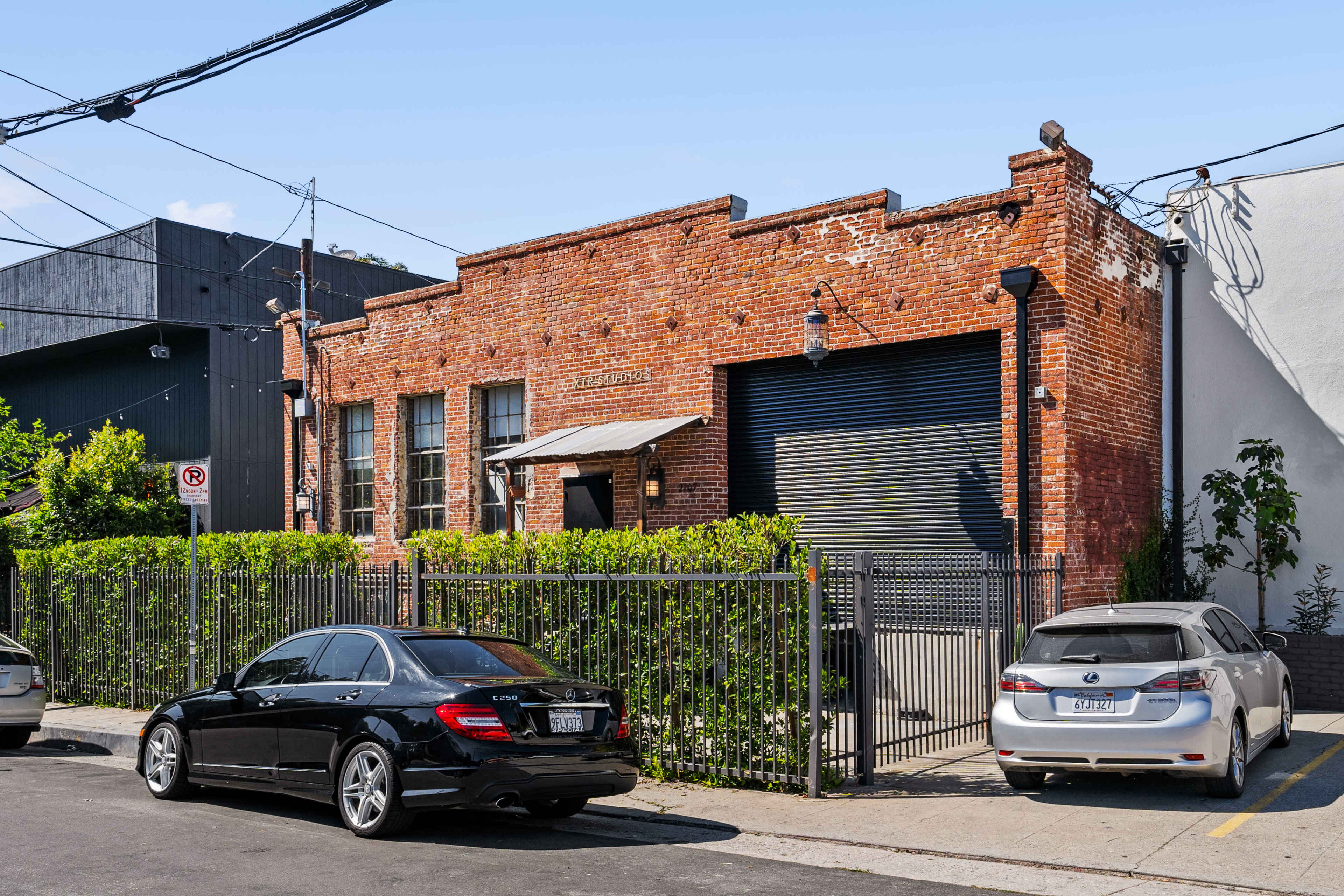 A brick building with a large garage door, bordered by a green hedge and parked cars in front.