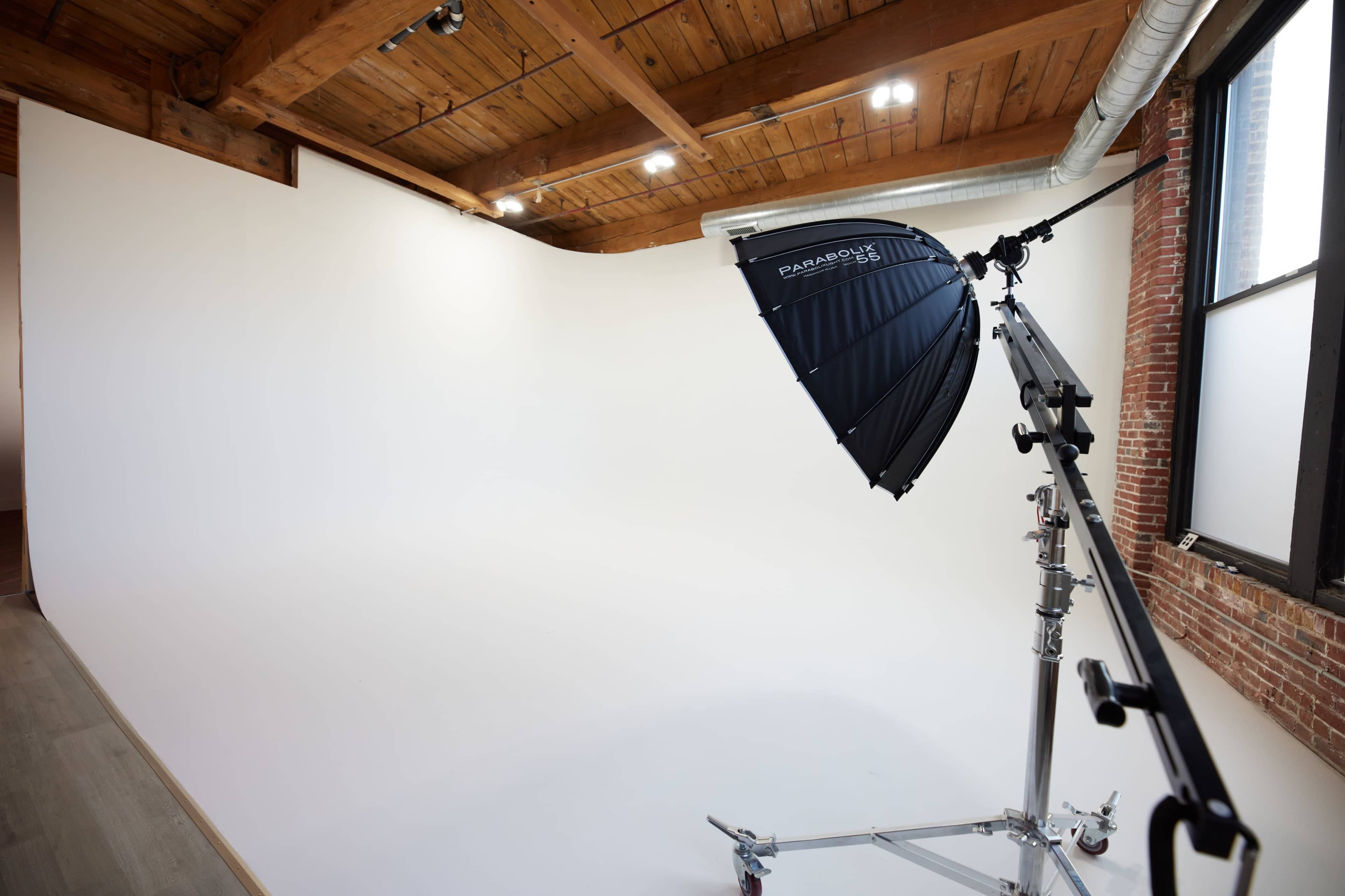 The image shows a photography studio with a white backdrop and a large umbrella light on a stand.