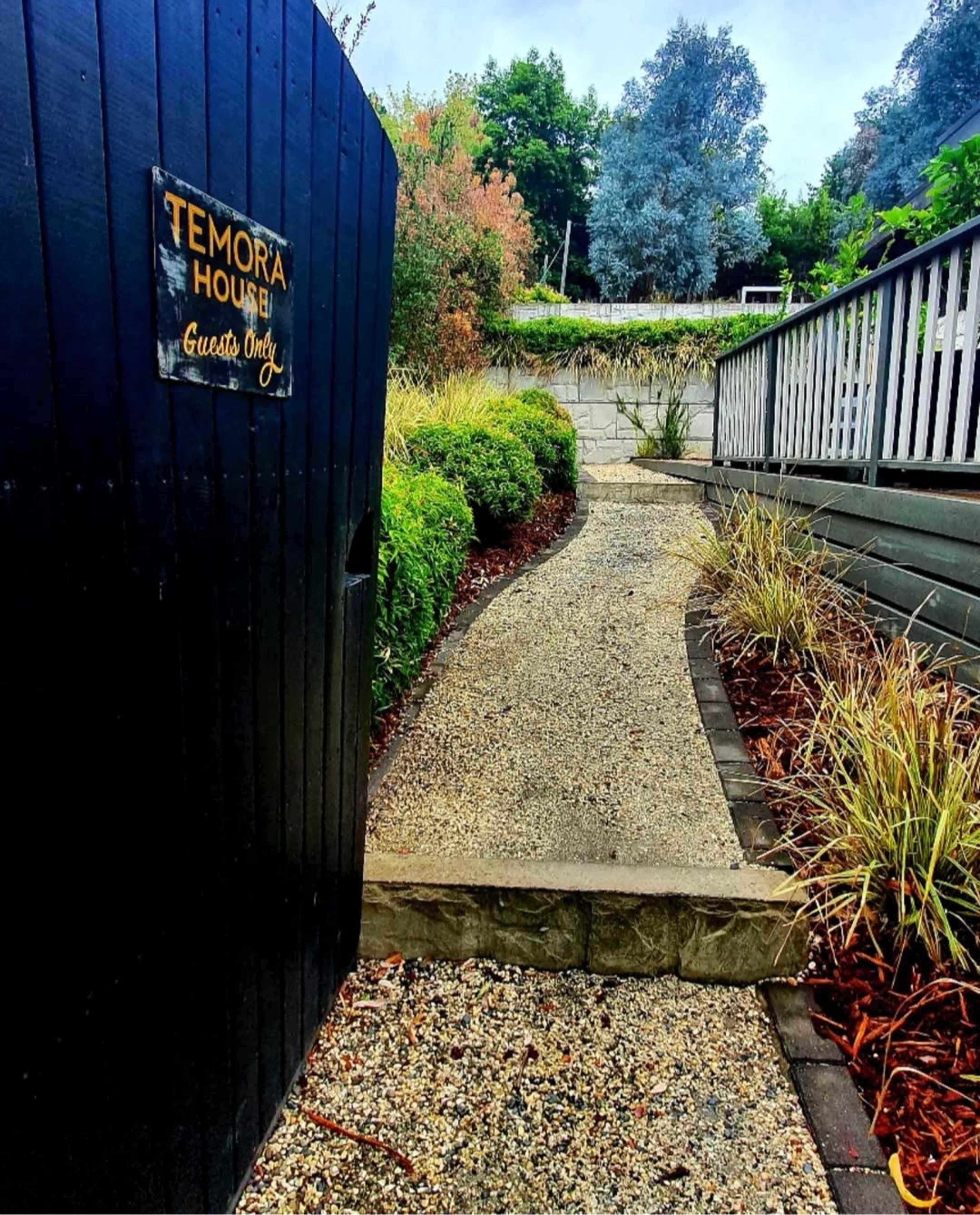 A gravel path leading through a garden gate marked "Temora House," leading to a wooden deck surrounded by greenery.