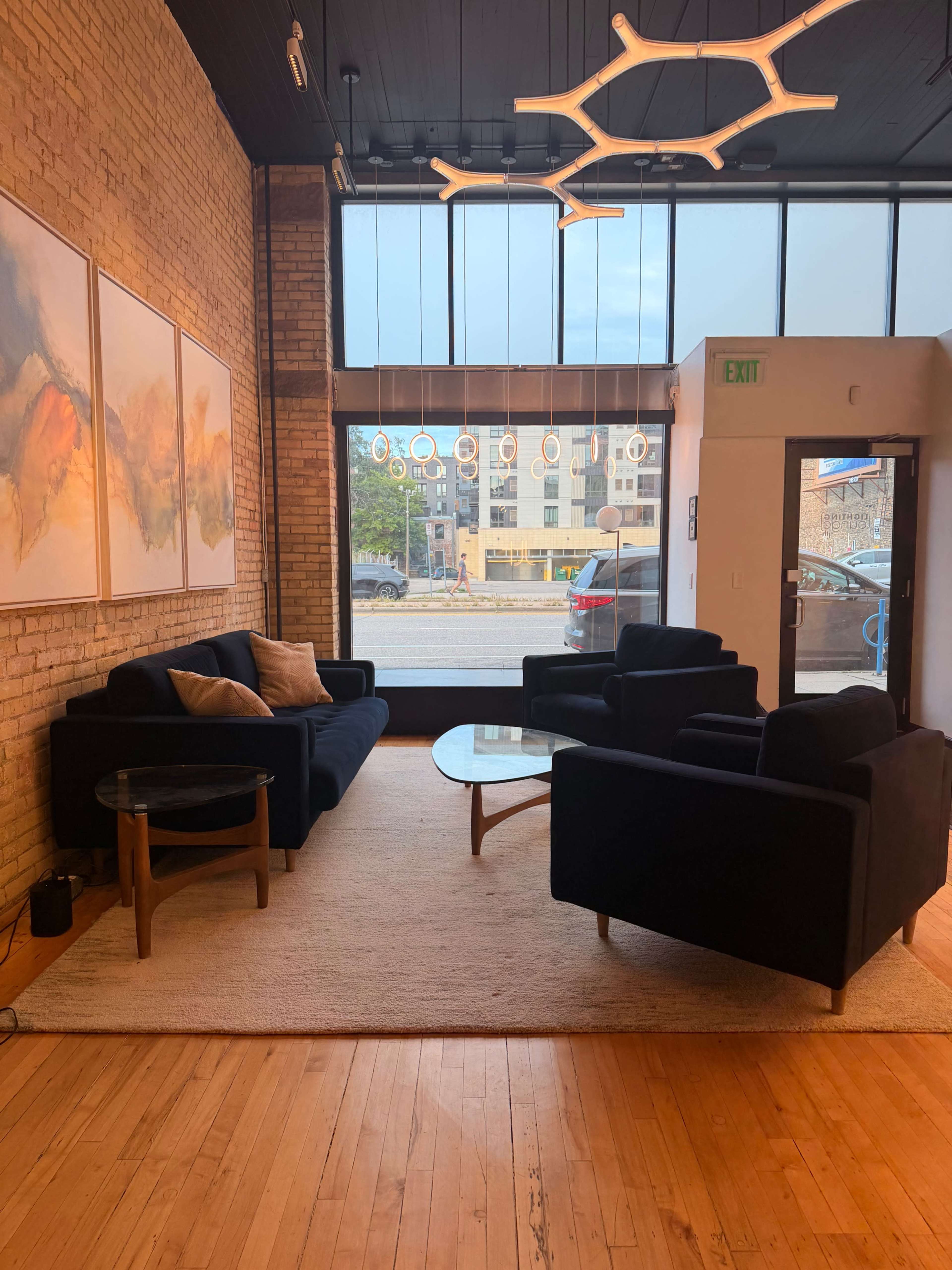 The image shows a modern interior space with two black sofas and a glass coffee table on a light-colored rug, illuminated by natural light from large windows.