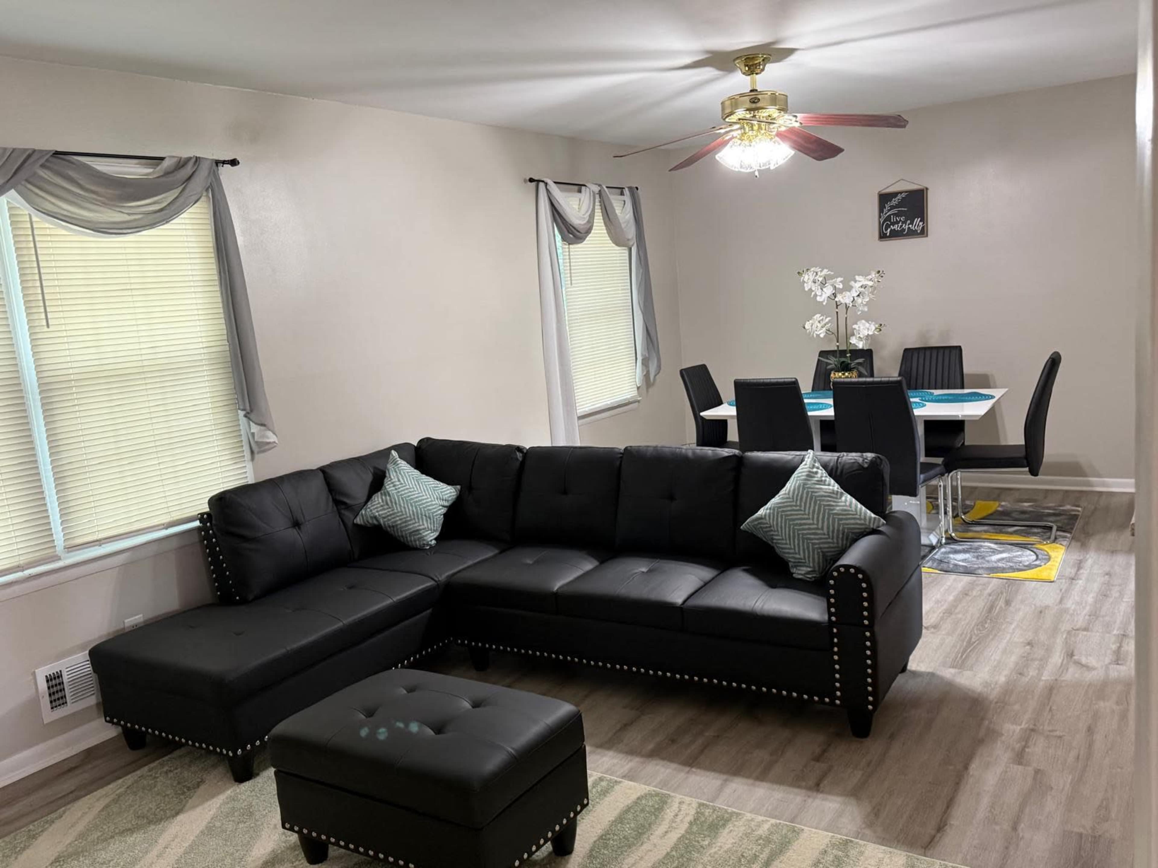 The image shows a living room featuring a black sectional sofa with decorative pillows, a matching ottoman, and a dining area with a table and chairs in the background.