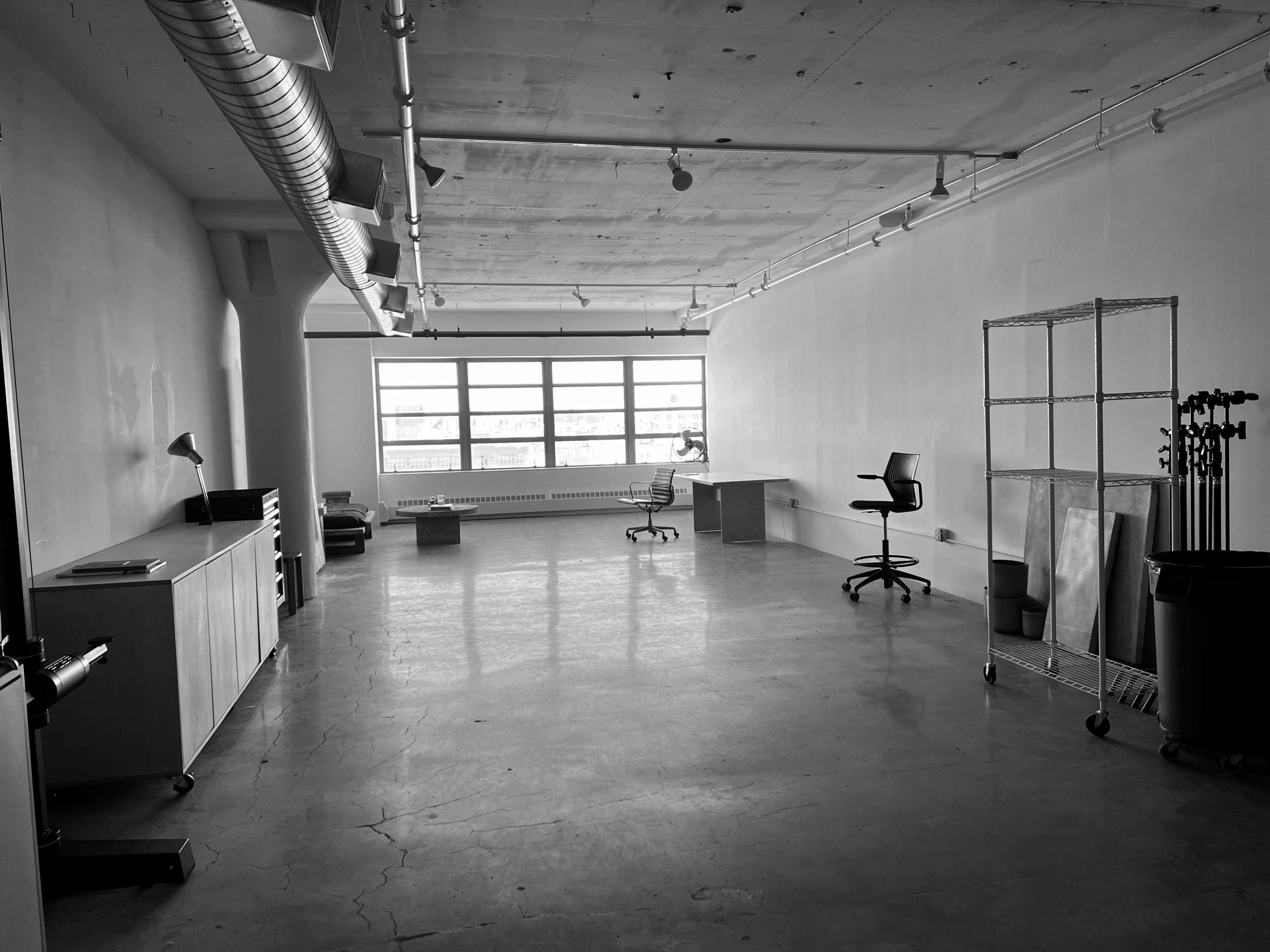 A large, empty room with concrete flooring, large windows on one side, and various pieces of furniture arranged sparsely, including a desk, chairs, and a shelving unit.