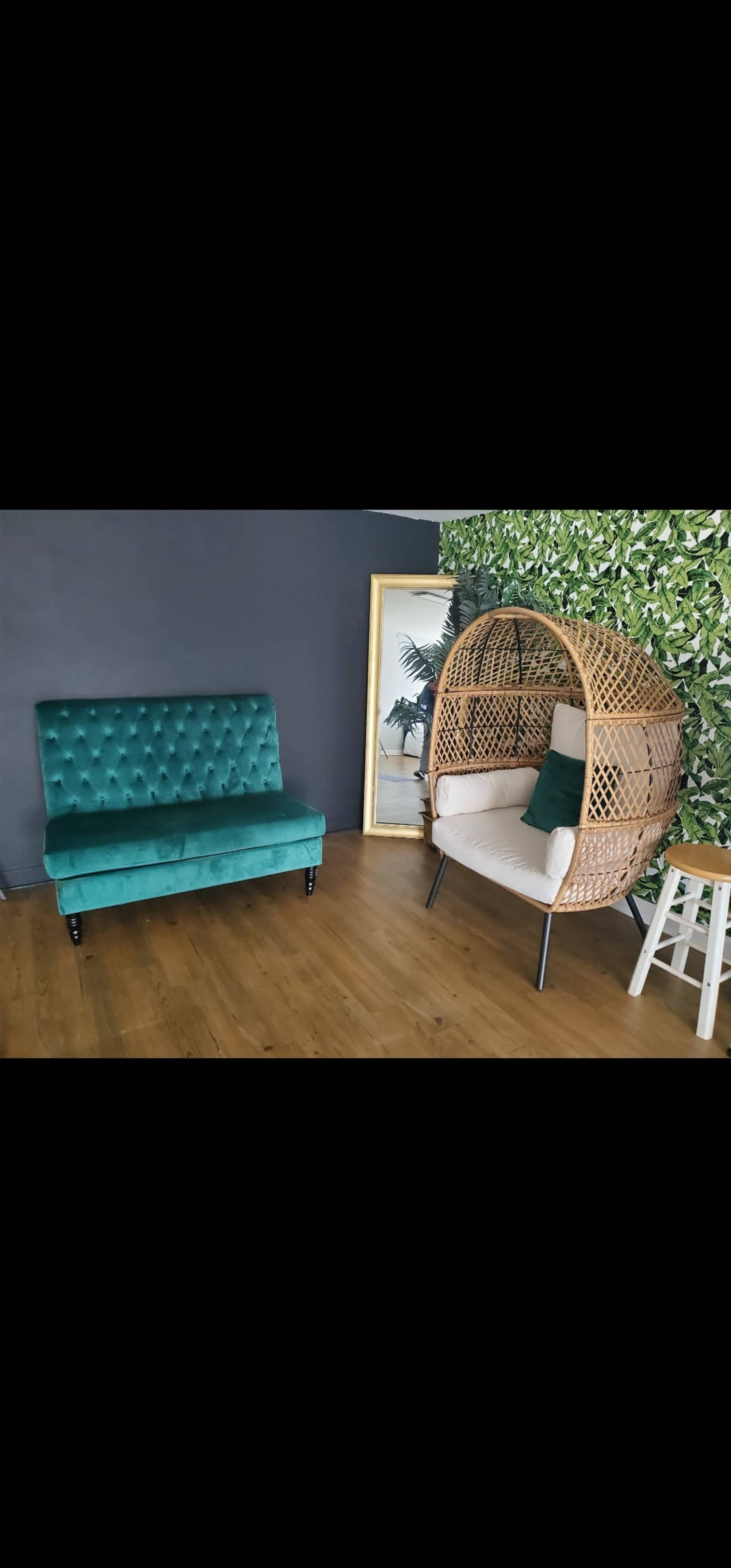 A stylish indoor setting featuring a green velvet sofa, a wicker hanging chair, a white stool, and a leafy wall backdrop.