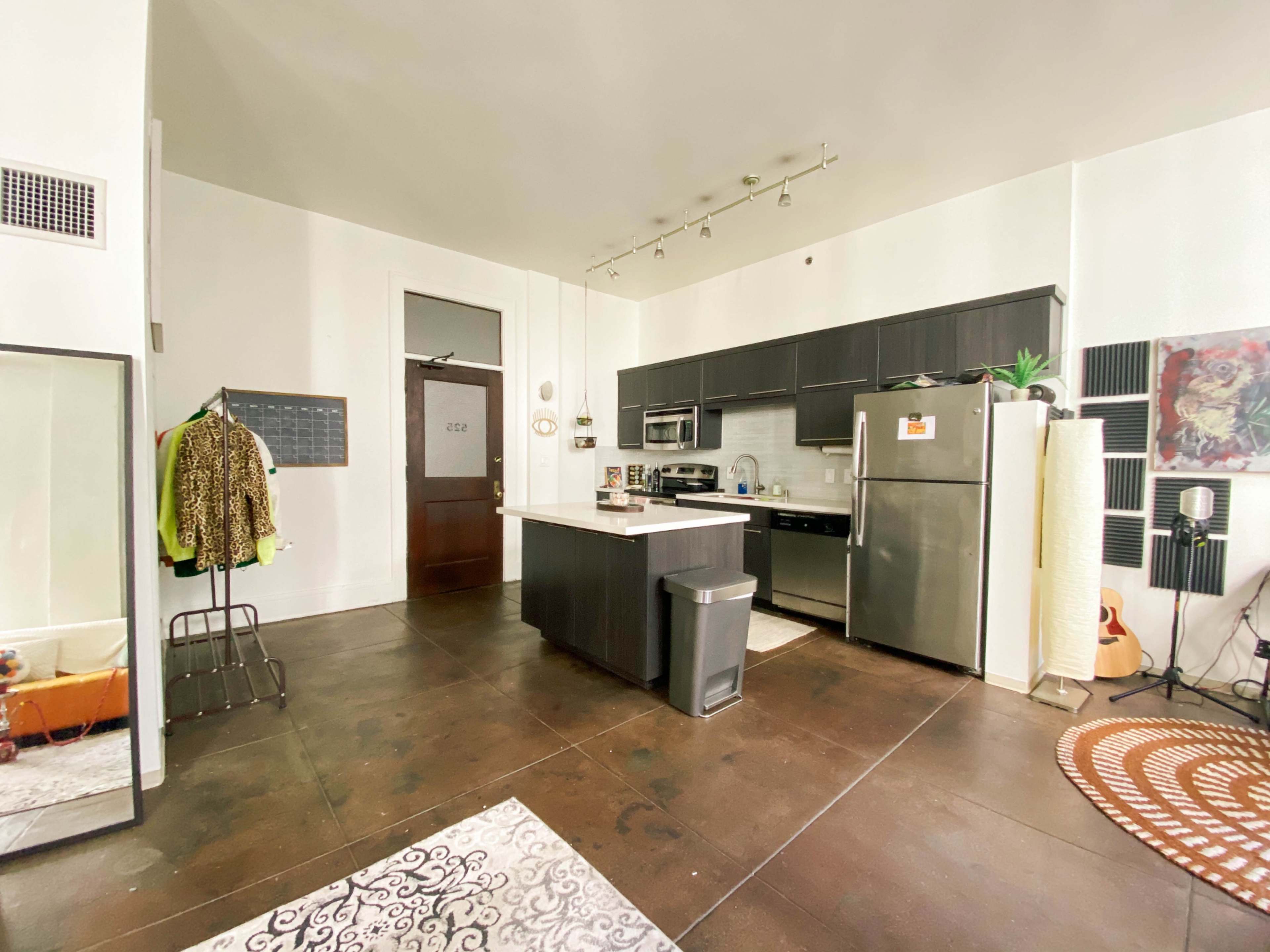 A modern kitchen with gray cabinetry, stainless steel appliances, and an open living space featuring a mirror, a clothing rack, and decorative rugs.