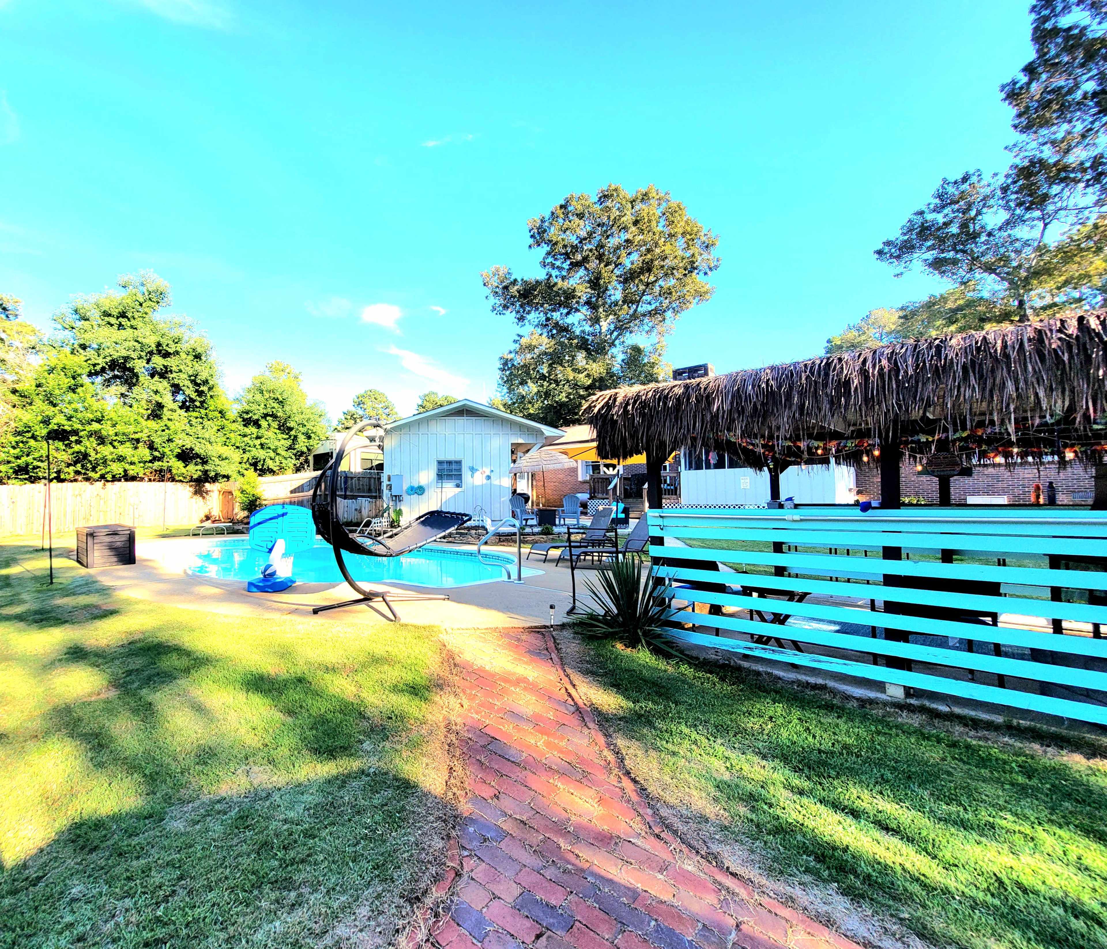 A backyard features a swimming pool surrounded by a patio, a tiki-style gazebo, and a hammock.