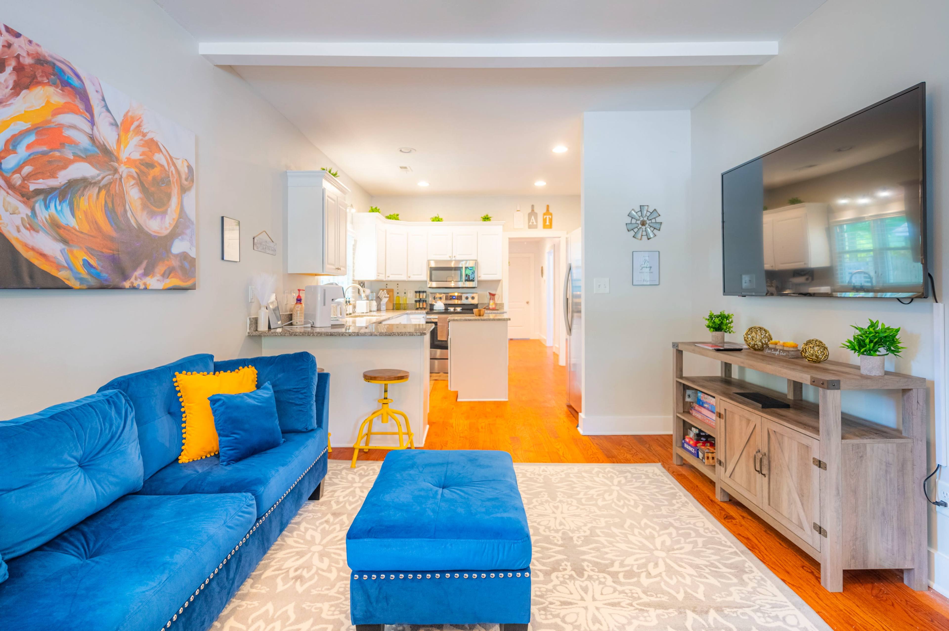 A bright living room with a blue sectional sofa, an ottoman, a television mounted on the wall, and a view into a modern kitchen with white cabinets.