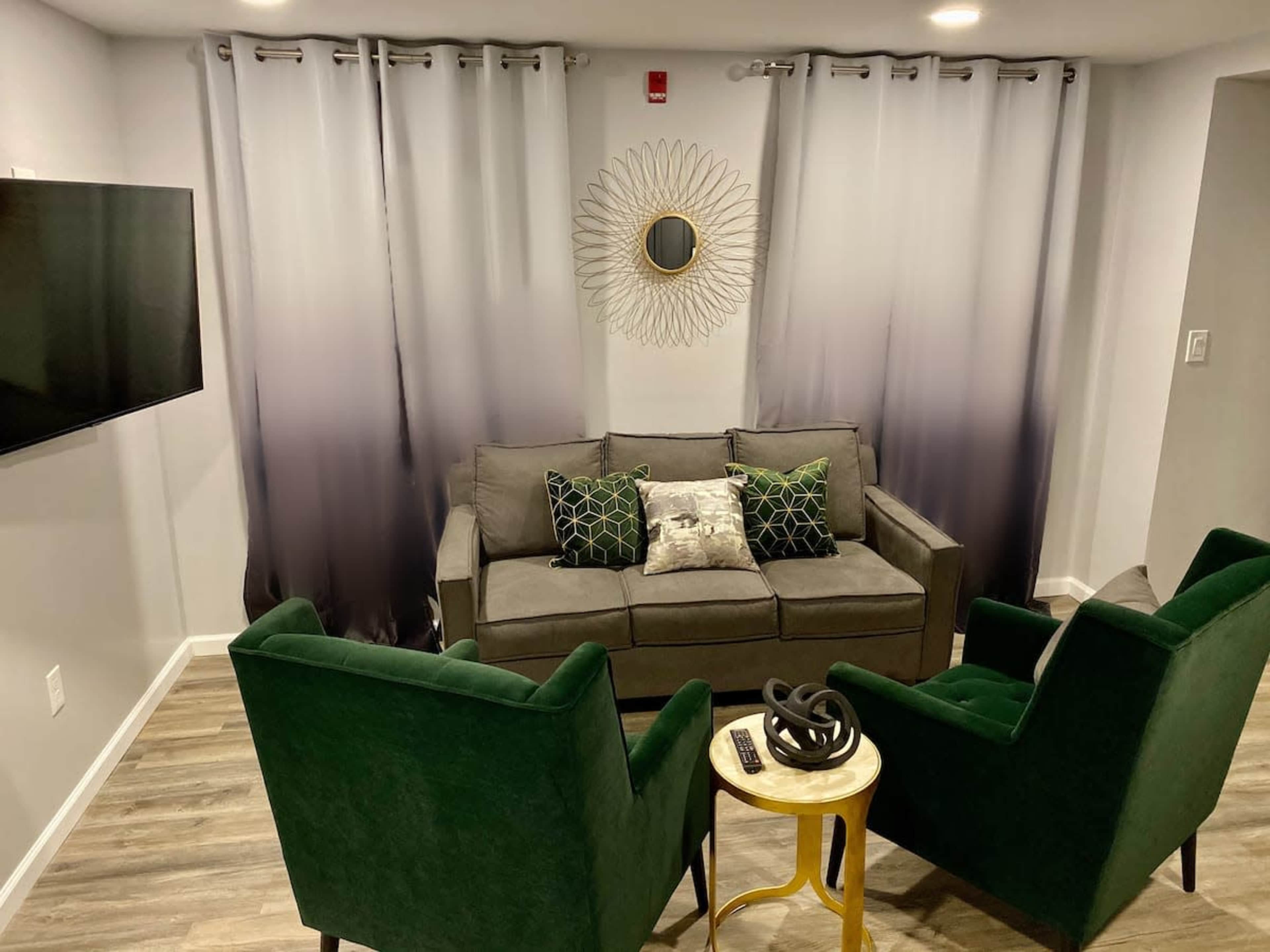 A modern living room featuring a gray sofa with decorative pillows, two green armchairs, a round wall mirror, and a small gold side table.