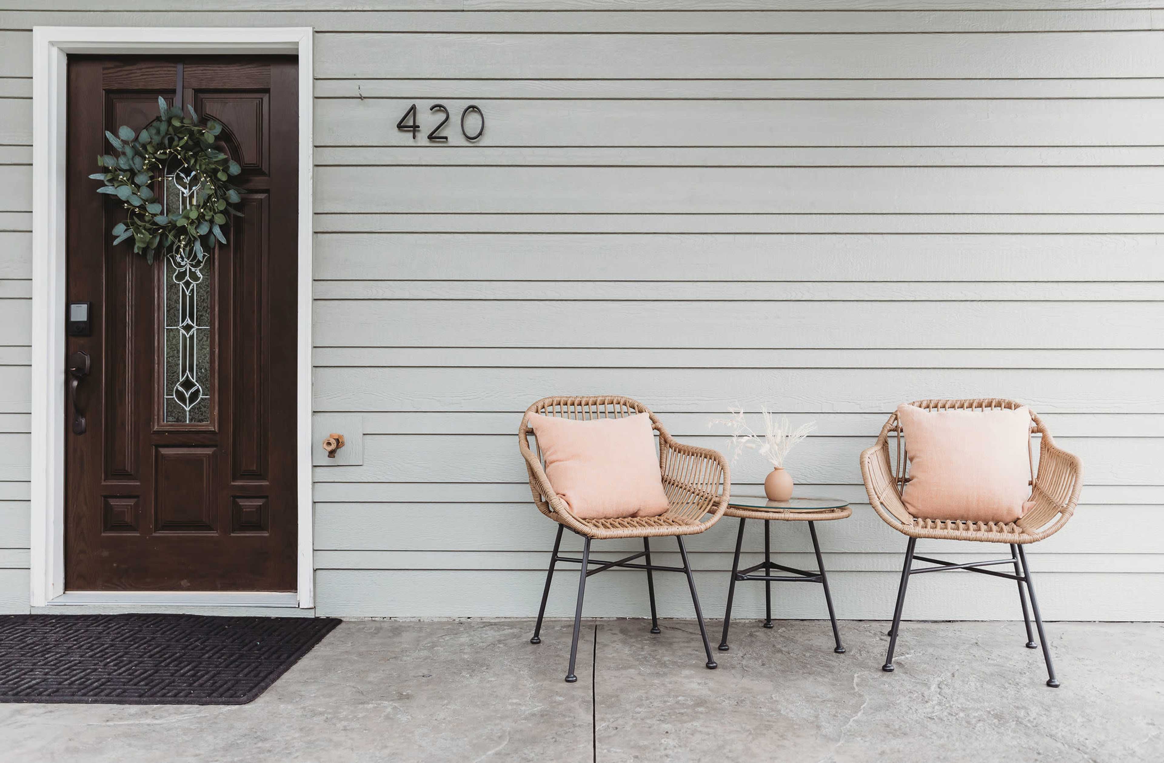 A front porch with a brown door, a decorative wreath, and two wicker chairs with cushions arranged around a small table.