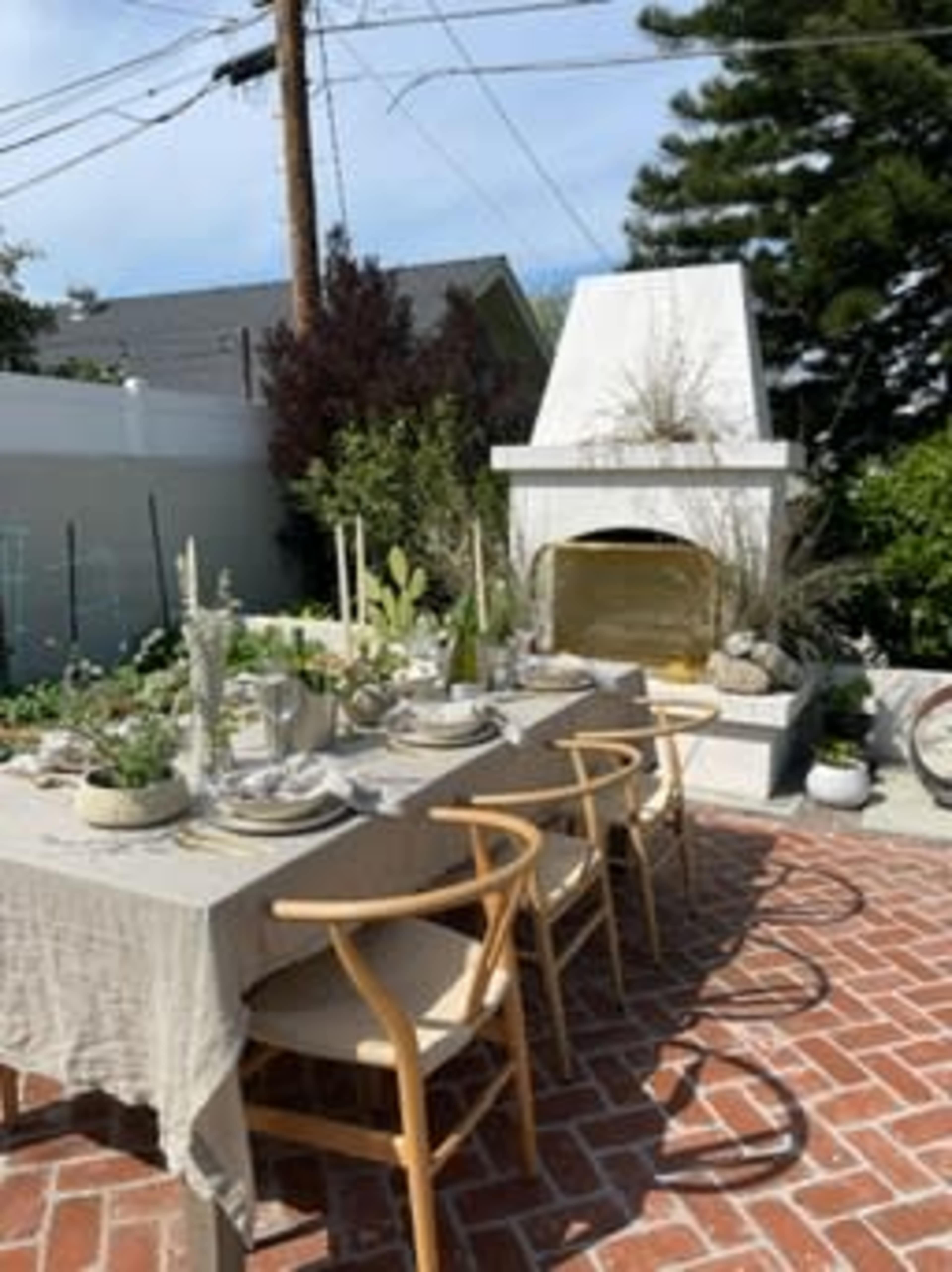 A long table set with dishes and plants is positioned on a brick patio next to an outdoor fireplace, surrounded by greenery and residential buildings.