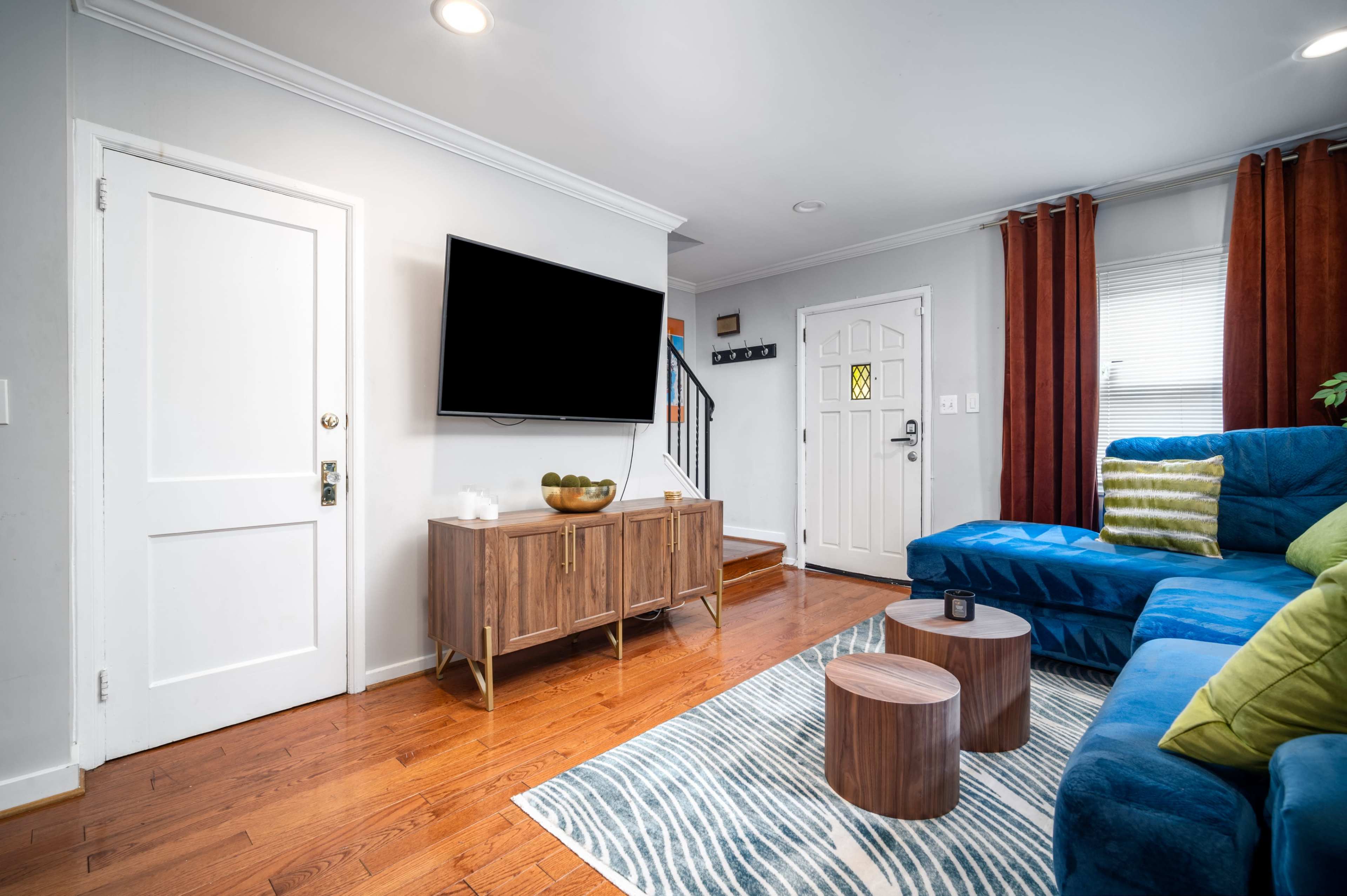 The living room features a blue sectional sofa, a wooden TV stand with decor, and large windows covered with dark curtains, all set against hardwood flooring.