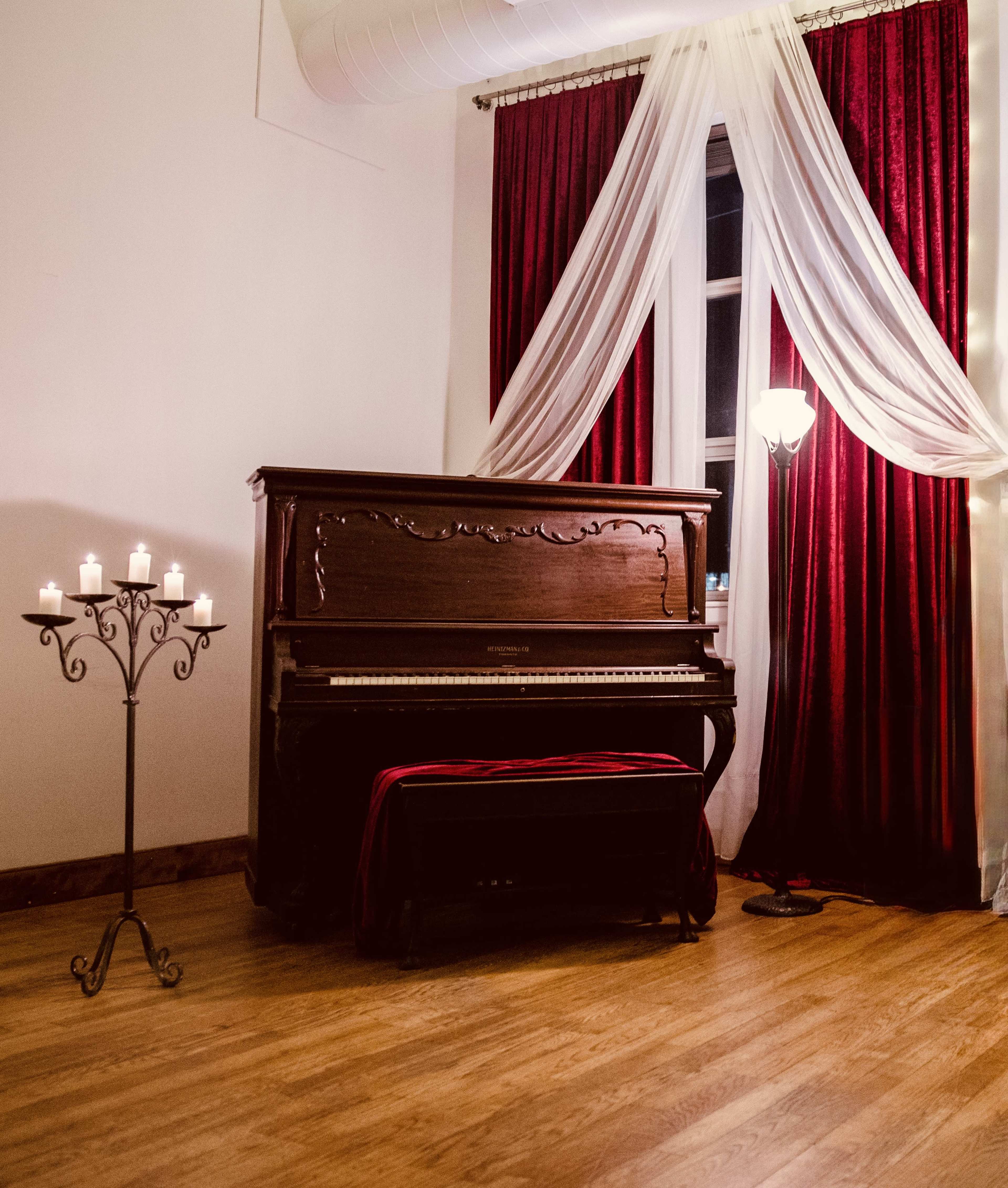 A vintage piano is placed against a wall, accompanied by a candle holder with several lit candles and draped curtains in red and white.