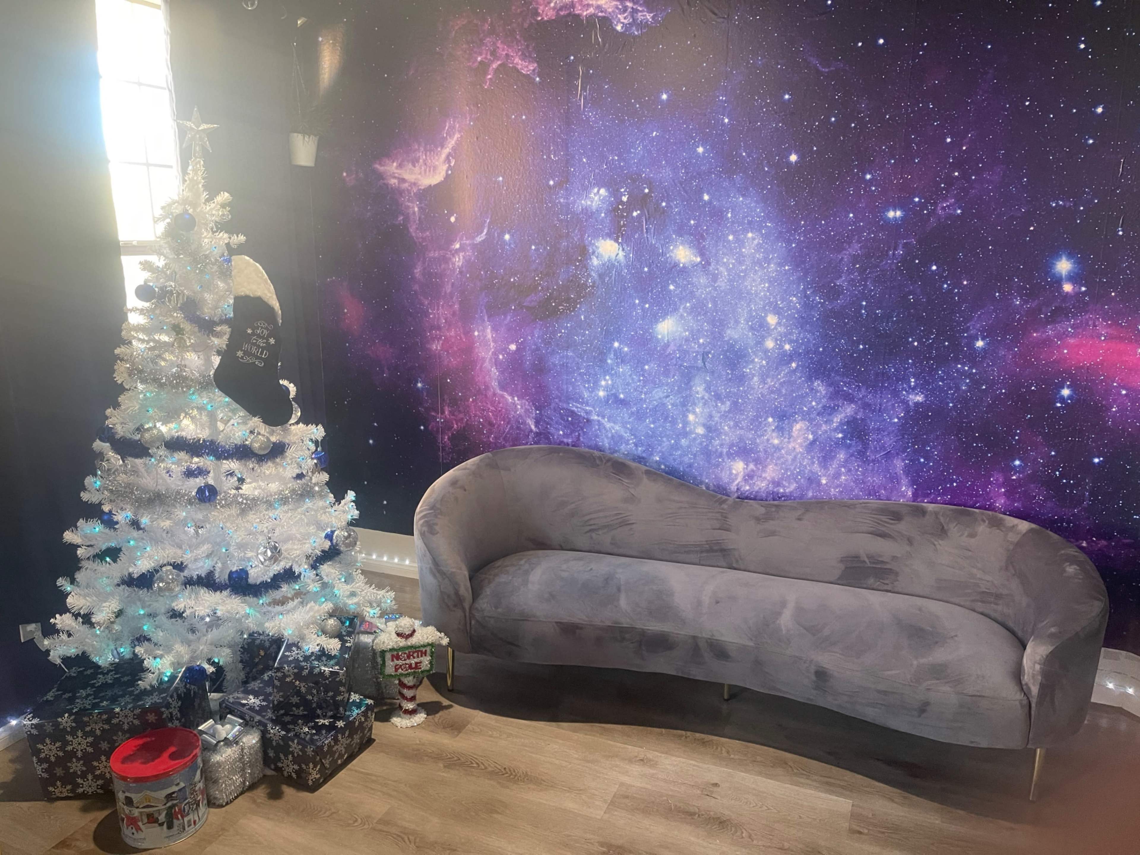 A Christmas tree decorated in white and blue lights is positioned next to a gray couch in front of a purple and pink galaxy-themed wall.